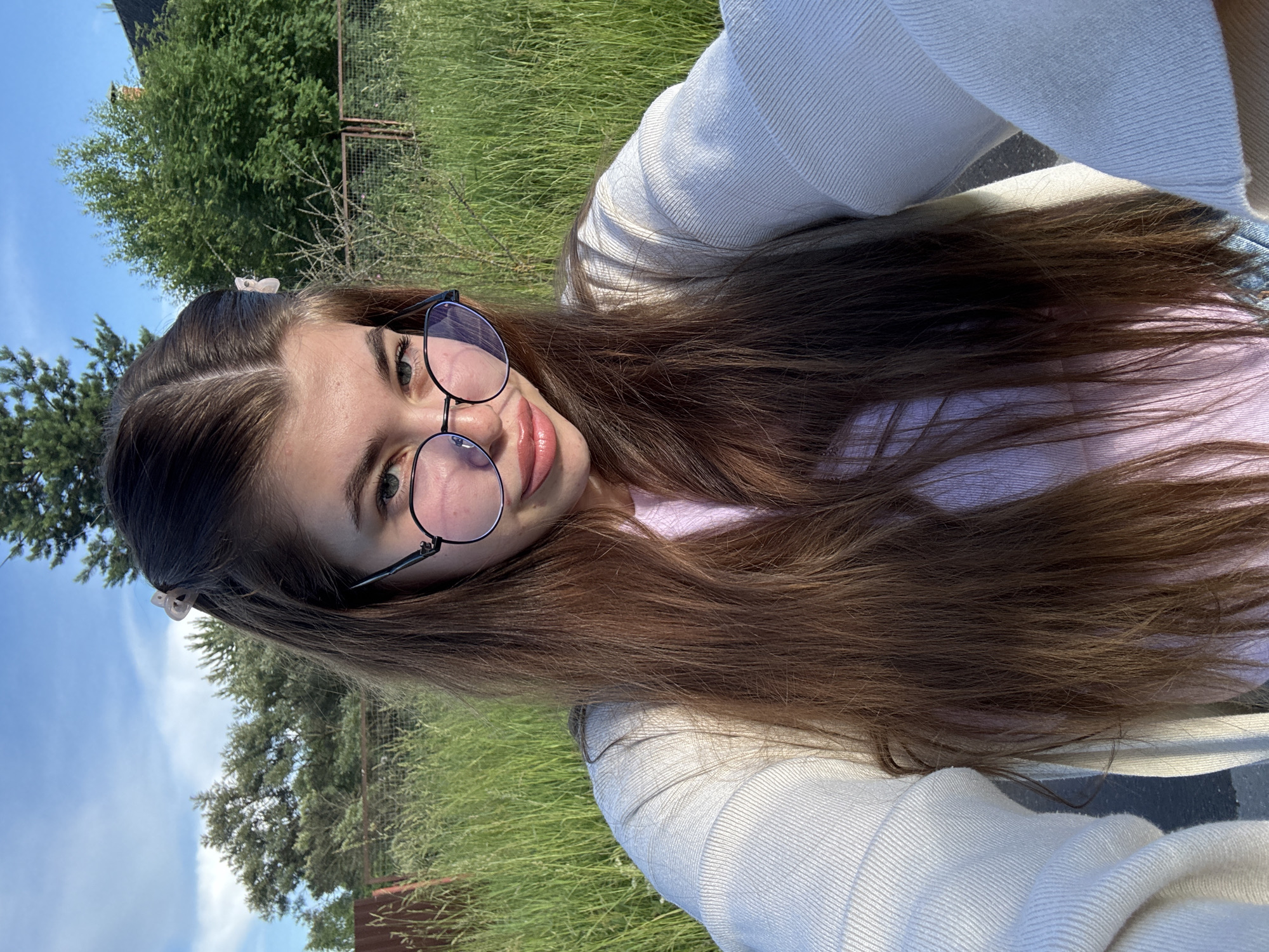 A young woman with long, brown hair and glasses is taking a selfie outdoors. She is wearing a light-colored sweater over a pink top. Her hair is partially visible, framing her face and falling down her chest. She has light-colored eyes and her lips are slightly parted. The background consists of tall green grass and trees, with a blue sky and some clouds visible. There is also a hint of a fence in the background. The lighting suggests it is daytime, possibly late afternoon given the warm tones. The overall scene appears casual and relaxed. The image seems to have been taken in a rural or park-like setting. There is no visible text in the image.
