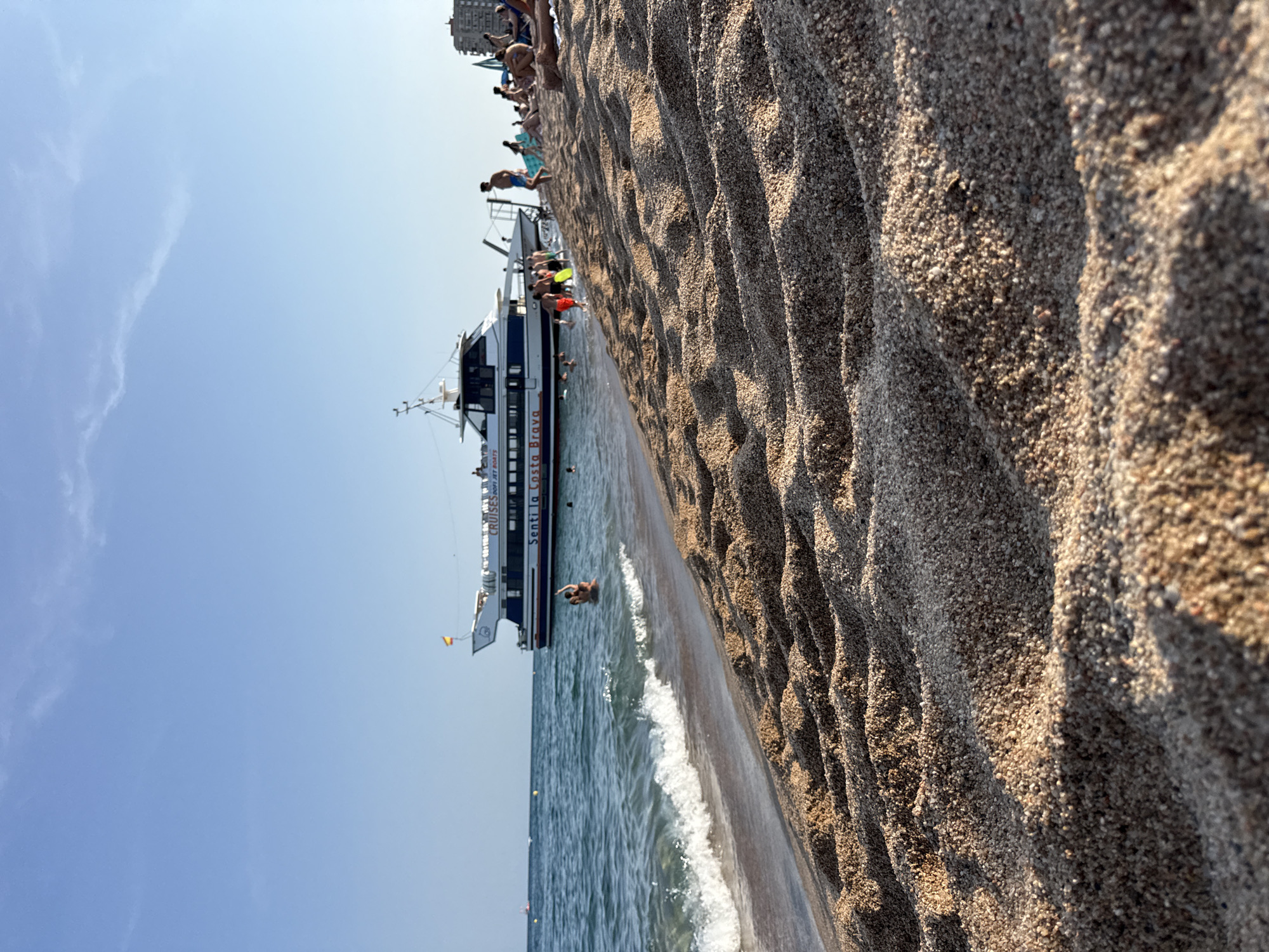 The image depicts a sunny day at the beach in Blanes, Spain. In the foreground, the textured sand of the beach slopes down towards the water. The sea is calm with small waves breaking near the shore. A white and blue passenger boat, named "Senti la Costa Brava," is anchored offshore. Several people are visible on the beach, some sunbathing and others in the water. The sky is clear blue with a few faint clouds. The scene conveys a typical summer day at a coastal resort.