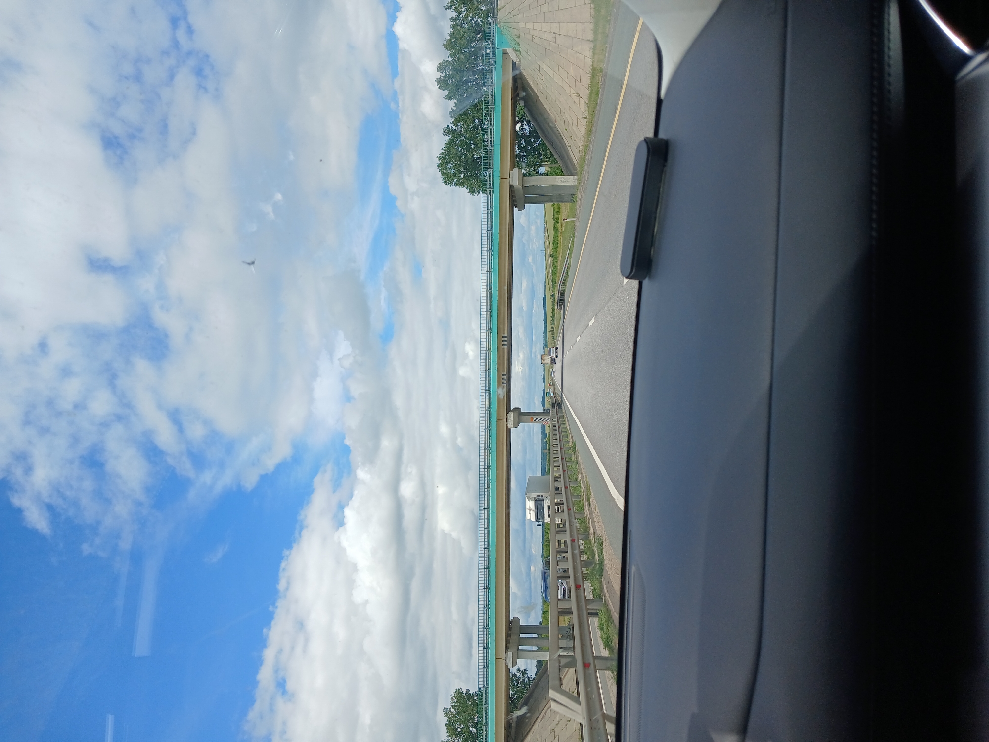 The image is a point-of-view shot from inside a vehicle, looking forward at a highway. A pedestrian overpass spans the highway, supported by concrete pillars. The sky is blue with scattered white clouds, and trees line the sides of the road in the distance. A white truck and other vehicles are visible on the highway, moving away from the viewer. The dashboard of the vehicle is in the foreground. The scene is a typical highway environment on a clear day. The location is identified as Rogatovo, Russia, based on the provided context, suggesting this is a highway or road within or near that area.