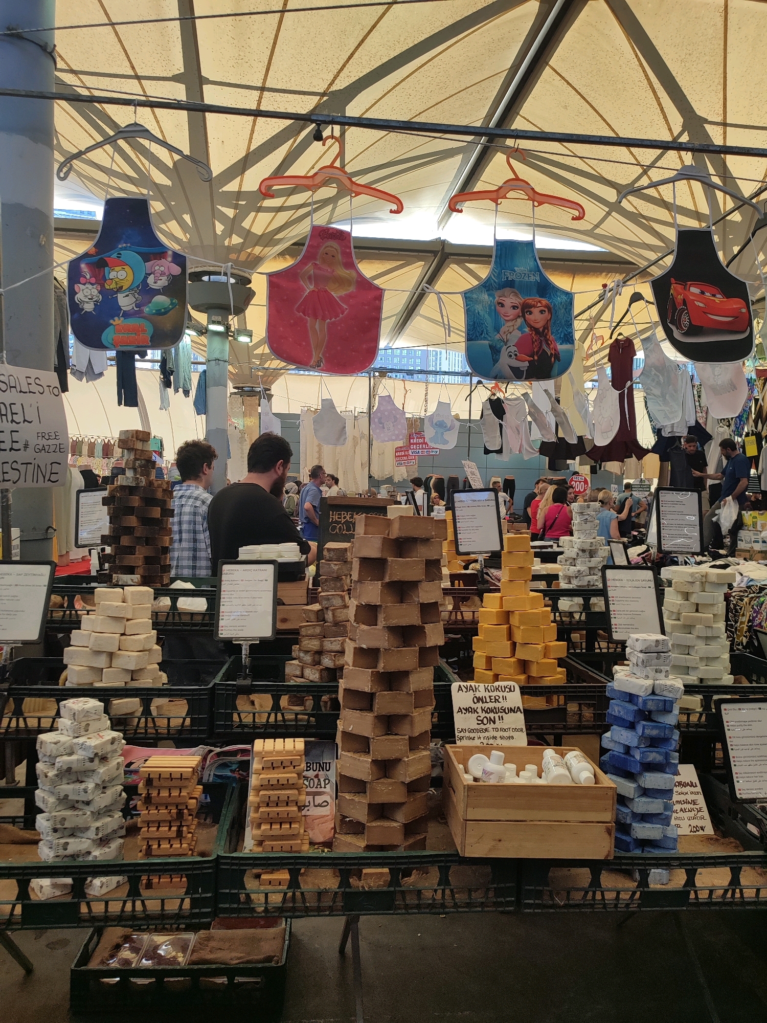 This image depicts a bustling market stall in Kadıköy, Turkey, likely within the Kadıköy Market area, known for its diverse offerings. The foreground is dominated by displays of handmade soaps in various shades of brown, yellow, and white, stacked in wooden crates and on shelves. One sign, partially visible, reads "BUNU SOAP" in English and Arabic script. In the mid-ground, several individuals are browsing or attending the stall. Two men are visible, one in a checkered shirt and another in a black t-shirt, seemingly engaged with customers or arranging merchandise. Behind them, a variety of colorful aprons, featuring cartoon characters like "Real Madrid," Barbie, "Frozen," and Lightning McQueen, are hung from hangers overhead. The market is covered by a cream-colored fabric canopy supported by a metal structure, creating a shaded environment. The background shows more stalls and shoppers, indicating a lively commercial atmosphere. The overall impression is of a vibrant, traditional market offering artisanal goods.
