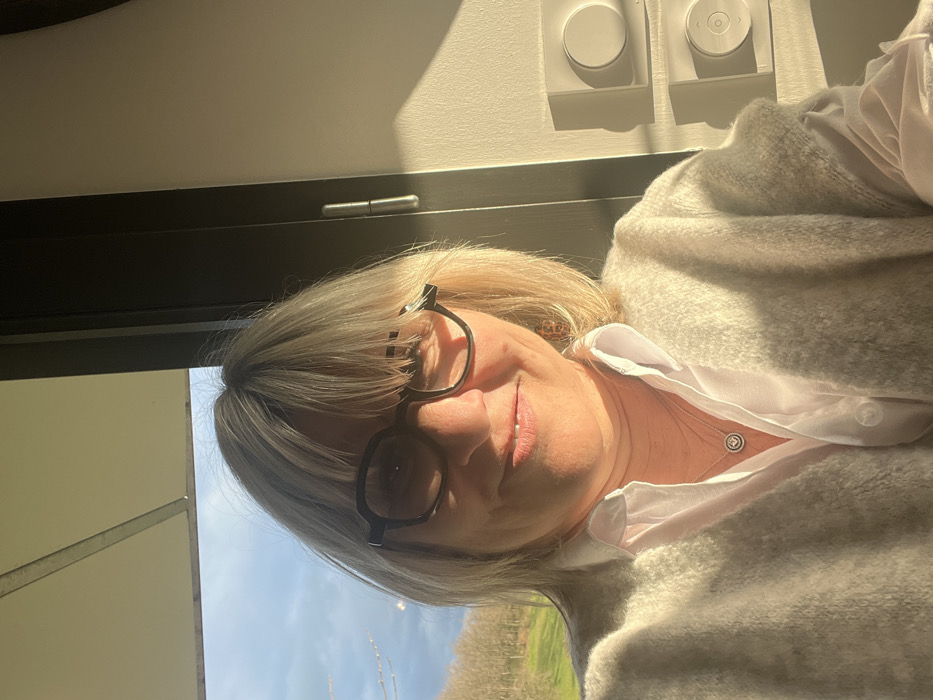 A blonde woman with bangs and dark-rimmed glasses smiles directly at the camera in what appears to be a selfie, located in Chambœuf, France. She wears a white collared shirt and a light-colored, possibly off-white, cardigan. A silver necklace with a circular pendant rests on her chest. Strong sunlight from the right illuminates her face and the interior space. To her left, a window reveals a clear blue sky with scattered clouds above a landscape of bare trees and green terrain. To her right, a light-colored interior wall features two white, rectangular wall-mounted devices, each with a circular control, casting shadows due to the direct sunlight. A dark vertical frame element, possibly part of a window or door, with a visible hinge, is situated between the woman and the wall devices.