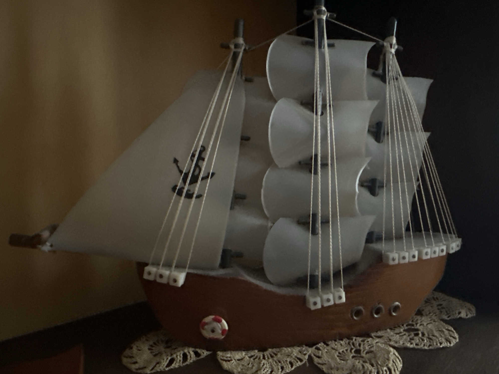 A model sailboat sits on a doily. The boat has a brown hull, white sails with a black anchor design on one, and white ropes. It is situated indoors, with a brown wall visible on the left and a dark surface on the right. The location is unspecified; the city of Troyan, Bulgaria is also not visible in the image.