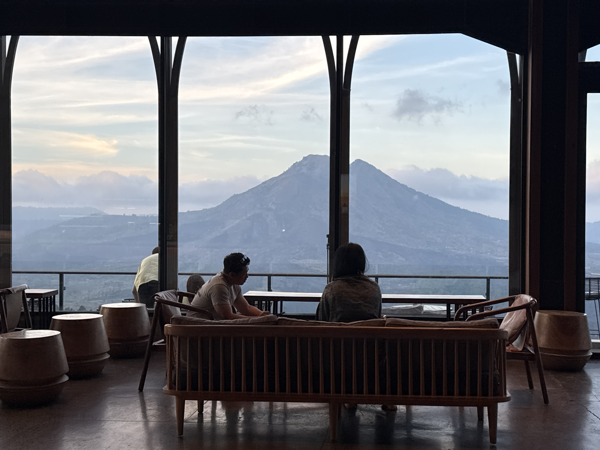This image depicts an indoor seating area with a panoramic view of Mount Batur in Kintamani, Indonesia. Two individuals are seated on a wooden sofa, facing away from the camera and towards the expansive mountain landscape. To the left, another person is standing, wearing a light-colored shirt, also looking out at the view. Several barrel-shaped wooden stools are placed in the foreground, along with chairs and tables. The setting appears to be a lounge or restaurant with large windows framing the volcanic scenery, which is bathed in the soft light of either dawn or dusk, with clouds visible around the mountain peaks. The overall atmosphere is serene and contemplative.