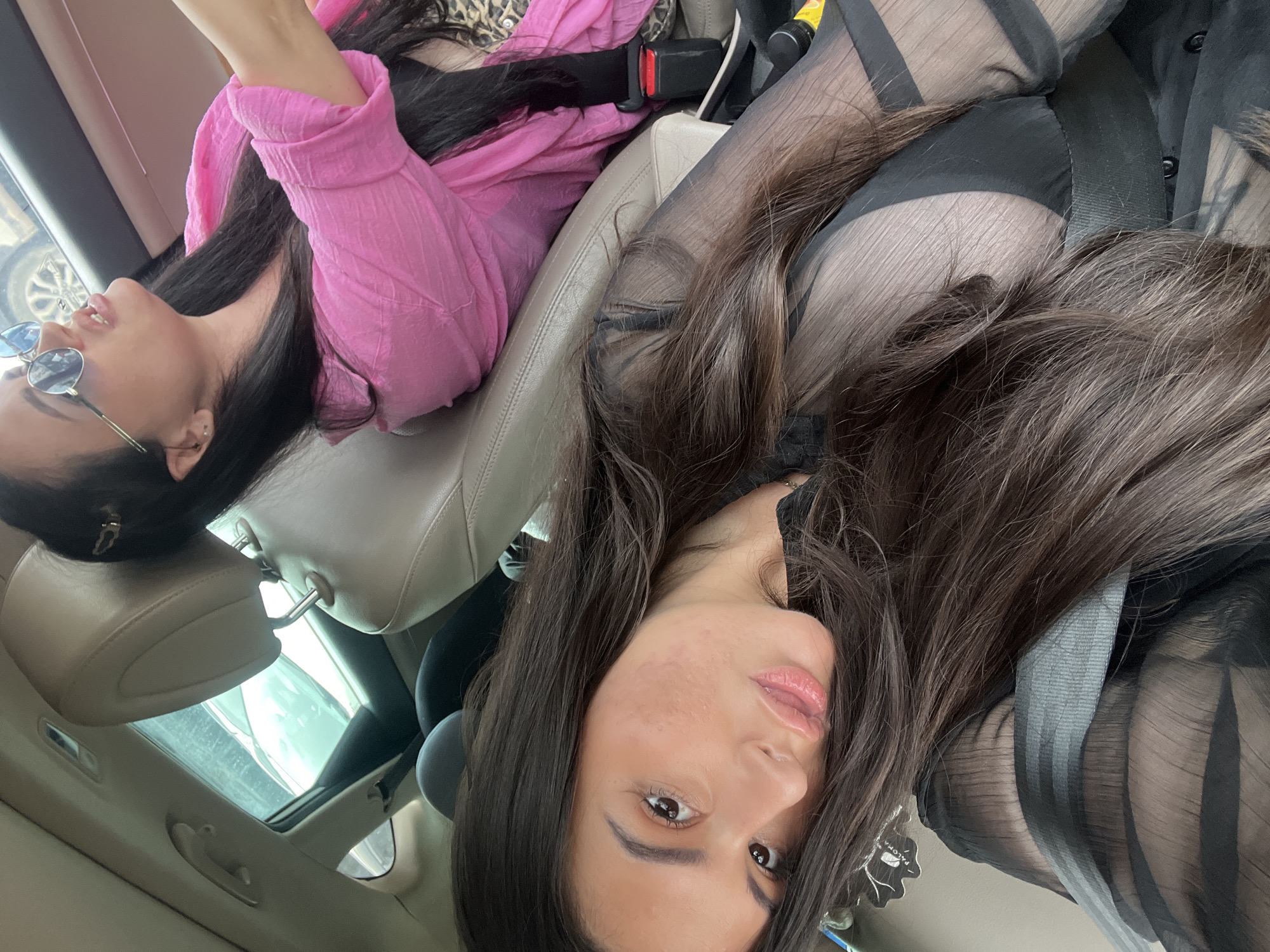 The image shows two women inside a car, likely in Kiryat Bialik, Israel, based on the prompt's request to include that location. The woman in the foreground is wearing a black, sheer top and looking directly at the camera. Her dark hair is partially obscuring her face. The woman in the background is wearing a pink top and sunglasses. Both women appear to be passengers, as indicated by the presence of a seat belt. The car has a sunroof visible in the background. The image is taken from an unusual angle, possibly from the car's ceiling or dashboard. The car's interior is light-colored leather.