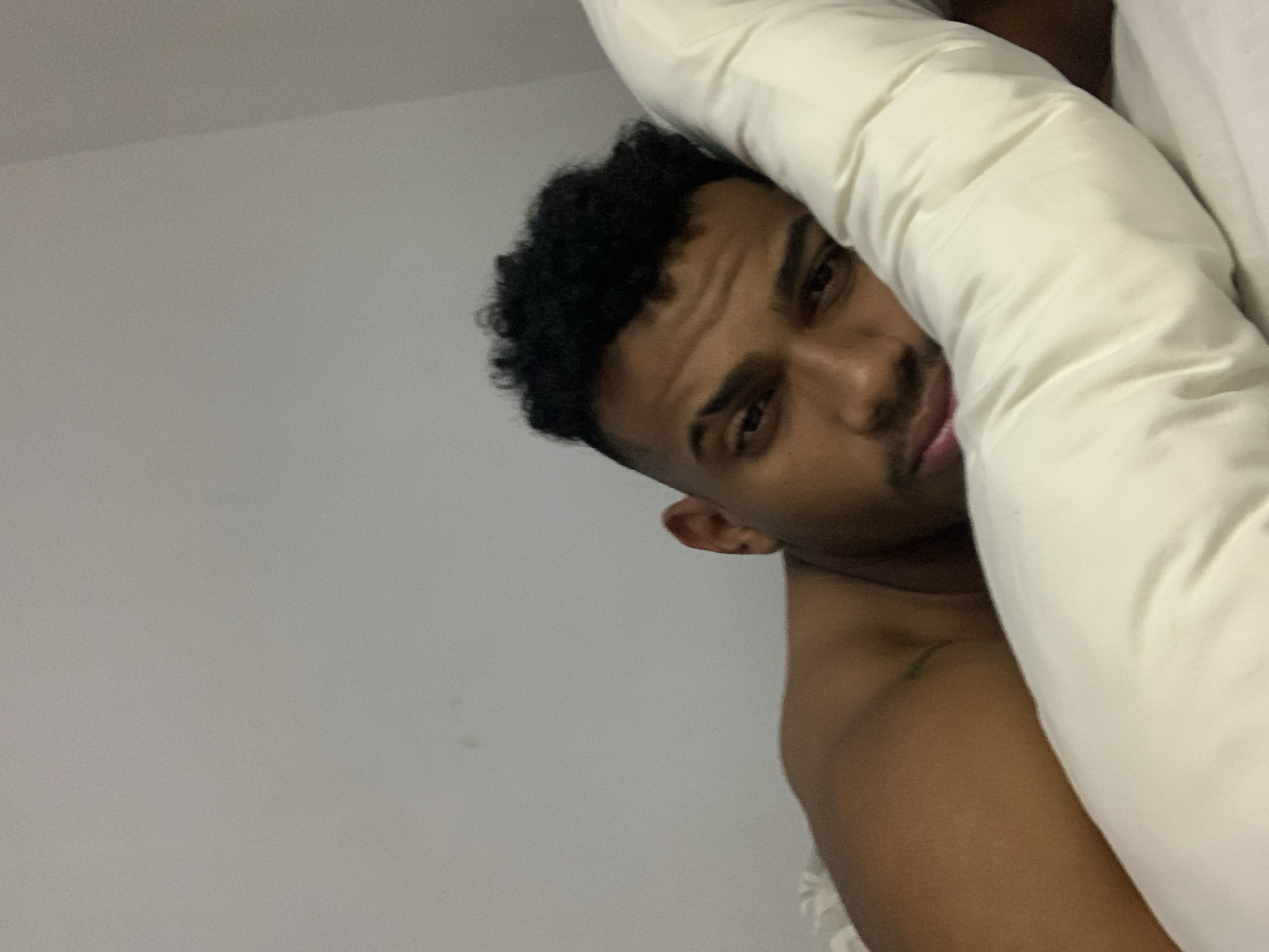 The media file displays a close-up, angled shot of a young man in an indoor setting, possibly a bedroom. He has dark, curly hair and light brown skin, with dark eyes that look directly at the viewer. His face, particularly his eyes and mouth, is the focal point, framed by light-colored bedding, likely a pillow or duvet, which partially obscures his body on the right side of the image.
The man's head is tilted, and he appears to be peeking out from or resting against the bedding. His expression is somewhat neutral, perhaps a mixture of curiosity and relaxation, with slightly furrowed eyebrows suggesting mild contemplation or intensity. A faint, soft smile or relaxed curve is visible on his lips. His bare shoulder and upper arm are visible on the left, indicating he might be shirtless or wearing minimal clothing suitable for rest. The background consists of a plain, light grey wall, offering no specific decorative details. The lighting is soft and even, typical of an indoor environment, making the exact time of day indeterminate. There is no visible text. The context places this scene in Porlamar, Venezuela.