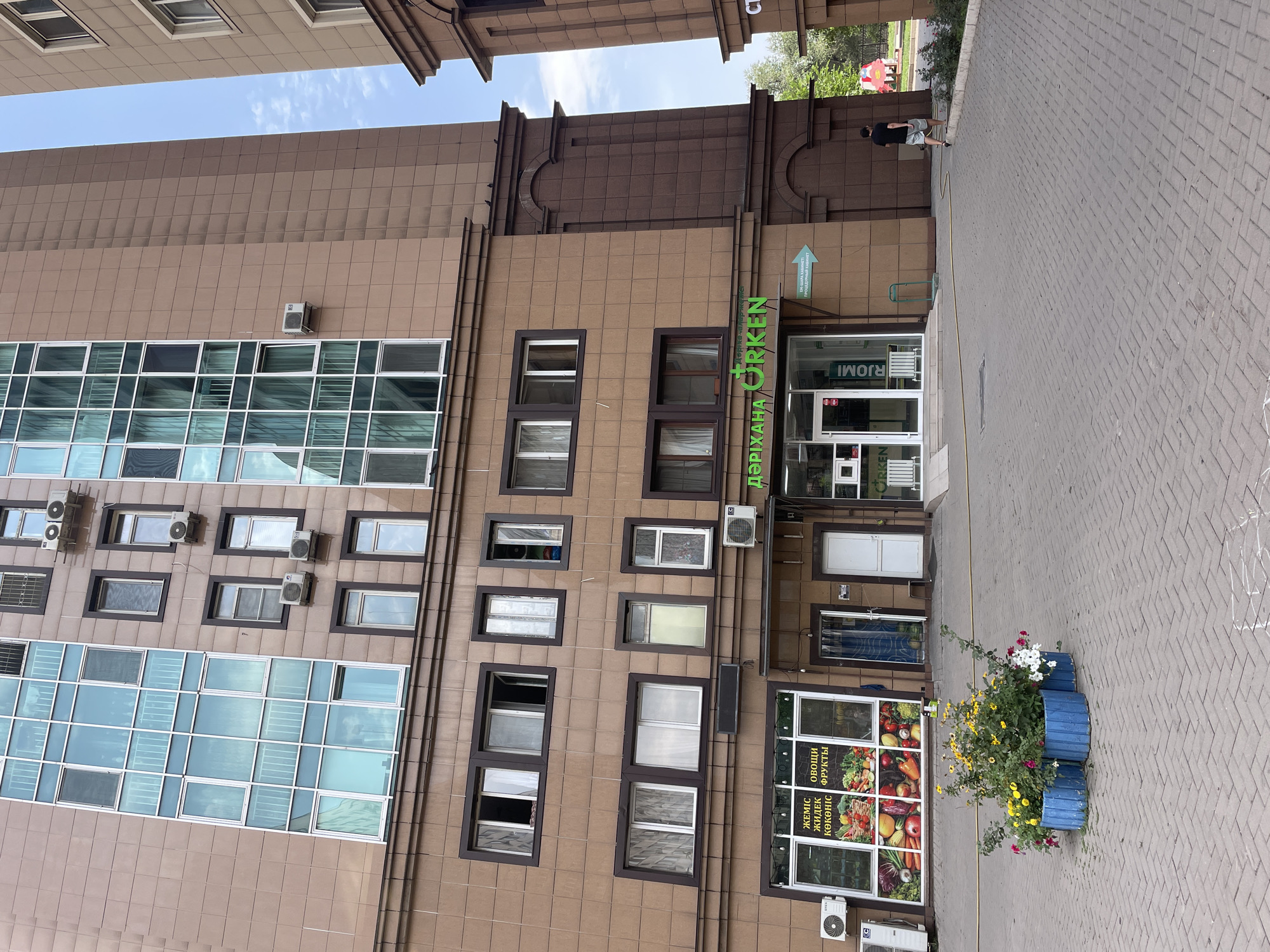 A man in a black t-shirt and grey shorts walks away from the camera on a paved area in front of a modern building in Almaty, Kazakhstan. The building has a brown facade and large windows. There are several shops on the ground floor, including one with a sign in Cyrillic that reads "DARIYAKHANA ORKEN" (Pharmacy Orken). A display of fruits and vegetables is visible in the window of one of the shops. In front of the building, there are three blue planters filled with colorful flowers. The sky is partly cloudy.