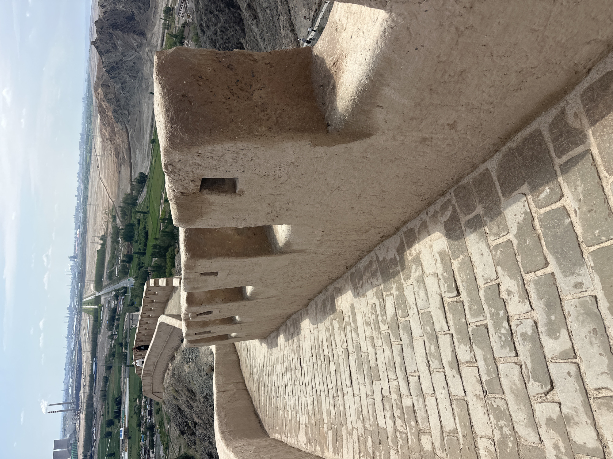 The image depicts a section of the Jiayuguan Great Wall in Jiayuguan, China, under a partly cloudy sky. The foreground is dominated by a winding pathway paved with weathered grey bricks, curving to the right. To the right of the path stands a section of the Great Wall constructed from earth and straw, with crenellations and small square openings visible along its top. The wall is a light brown, textured material. In the middle ground, a few individuals are walking along a slightly elevated portion of the wall. Beyond the wall, the landscape transitions to rolling hills and a valley. In the distance, a sprawling industrial area with smokestacks emitting plumes of white smoke is visible, along with a city skyline and power lines stretching across the terrain. The overall scene showcases the juxtaposition of historical architecture with modern industrial development.