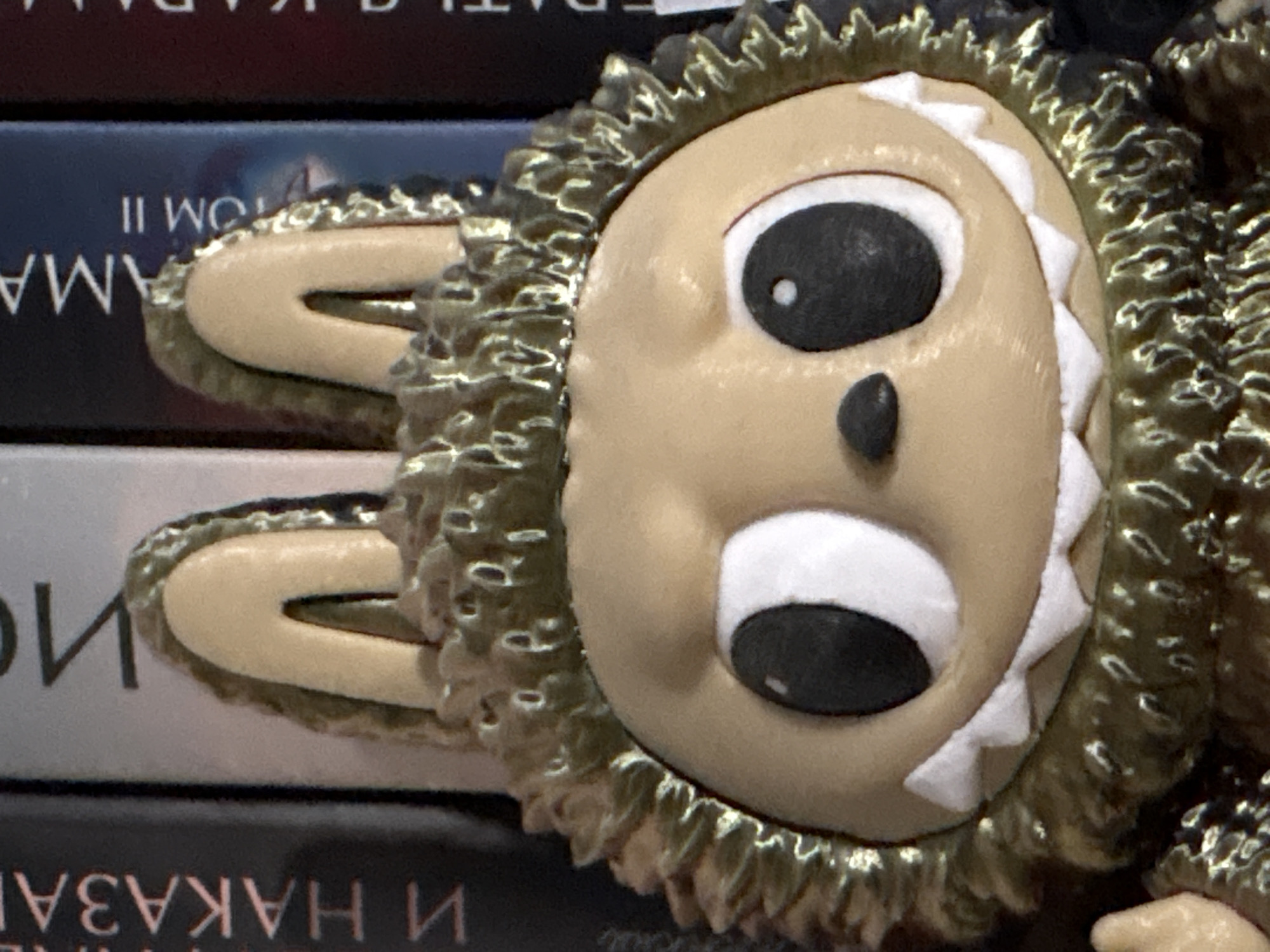 The image is a close-up of a doll or figurine with a cartoonish face featuring large black eyes with white highlights, a small black nose, and a wide open mouth. The face is light tan, with white jagged shapes around the mouth. The doll has two floppy, tan-colored ears that appear to be attached to its head. The face is surrounded by a textured, possibly golden or metallic material, possibly representing fur or hair. The doll is positioned against a stack of books. Visible text on the books appears to be in Russian, including "TOM II," "AMA," and partial words like "HAKAZA." There is no clear indication of Izhevsk, Russia.