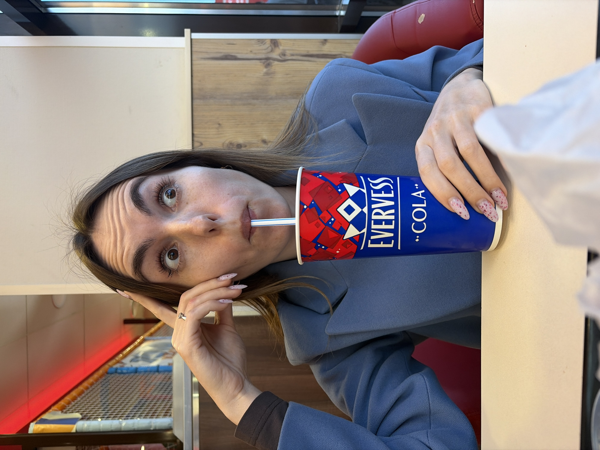 An adult female is depicted seated at a light-colored table within a commercial dining establishment in Izhevsk, Russia. She has medium-length brown hair and is wearing a blue-grey blazer over a dark long-sleeved undergarment. Her fingernails are manicured with light pink polish and specks of glitter. A silver ring with a clear stone is present on her left ring finger. Her facial expression shows wide-open eyes, directed slightly upward and to the right, with raised eyebrows and a furrowed forehead. She is actively consuming a beverage from a blue, red, and white paper cup labeled "EVERVESS -COLA-" via a white and blue striped straw. Her left hand is raised with the index finger touching her temple area, while her right hand holds the cup. A white crumpled paper object is partially visible on the table in the foreground, bottom right. The background features a children's play structure with orange and blue netting and a red illuminated overhead fixture. The walls are two-toned, with a light beige/off-white upper section and light wood paneling below. A red upholstered chair back is visible to her right. A window or glass door is visible on the far right, suggesting an exterior view. The environment is consistent with a fast-food restaurant or family-friendly cafe, likely featuring a children's play area, commonly found in urban settings like Izhevsk.