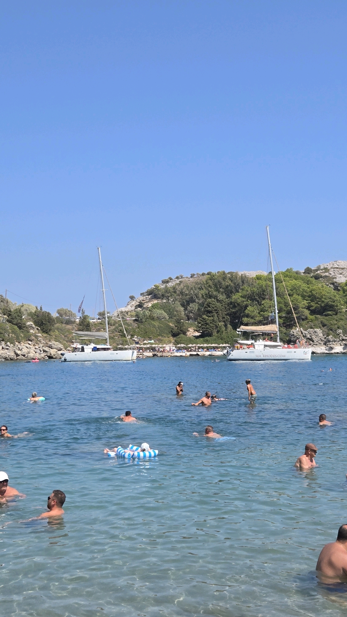 This is a vertical, outdoor, daytime photograph taken at the beach in Anthony Quinn Bay, Rhodes, Greece. The clear, blue water is filled with people swimming and floating on inflatable rings. Two sailboats are anchored in the bay, one larger than the other. In the background, a rocky hillside covered with lush green trees slopes down to the water's edge. Several beach umbrellas are visible along the shoreline. The sky is a bright, clear blue.