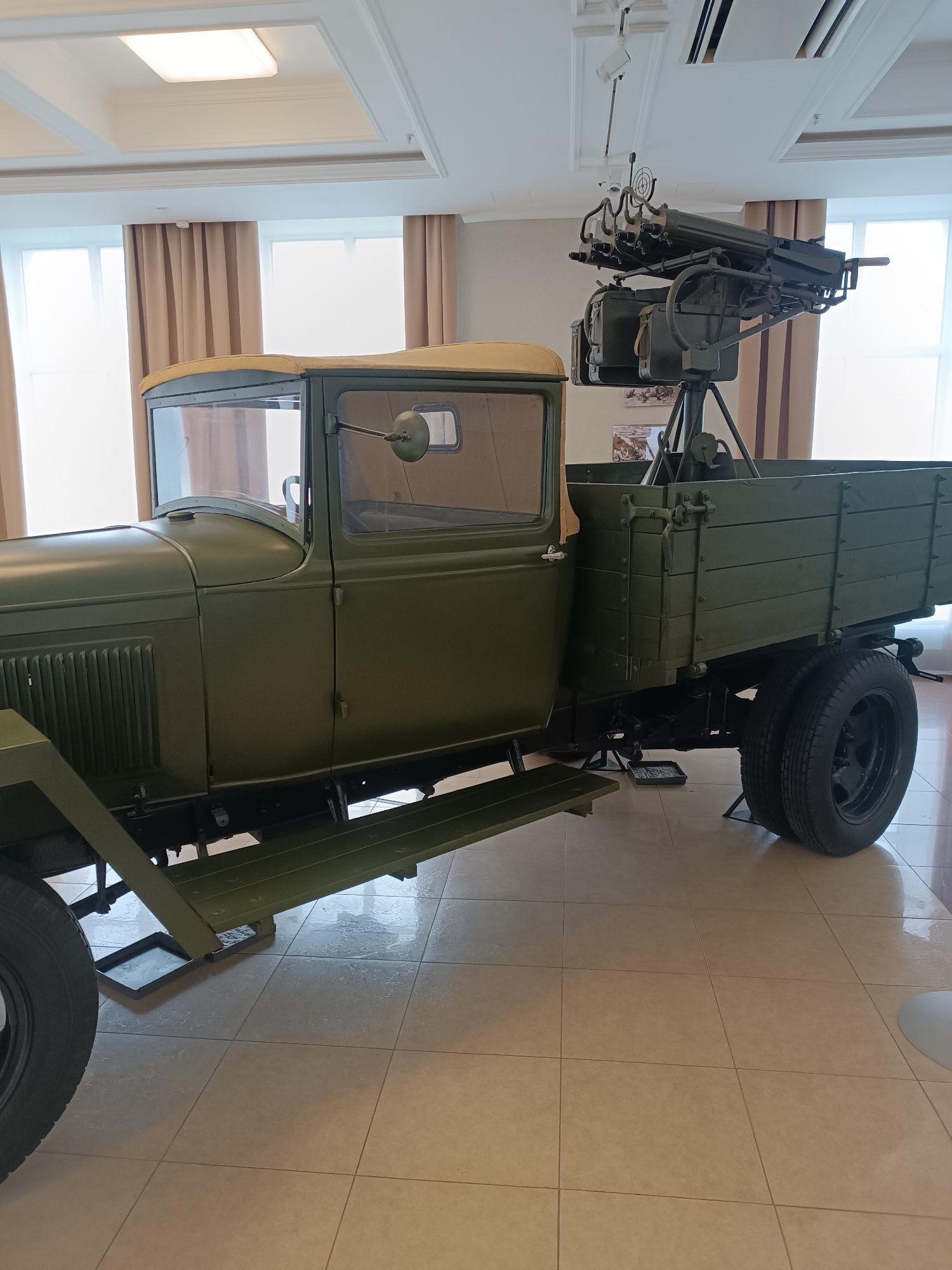 The image shows a vintage military truck with a mounted machine gun, displayed indoors. The truck is olive green, with a canvas roof over the cabin and wooden sides on the cargo bed. The machine gun is positioned on a tripod mount in the rear of the truck bed. The setting appears to be a museum, with light-colored tiled flooring and large windows with beige curtains in the background. Above the truck, a modern ceiling with recessed lighting fixtures is visible. The environment is clean and well-lit. This artifact is part of the military technology exhibition at the Museum of Military Technology "Battle Glory of the Urals" in Verkhnyaya Pyshma, Russia. No people are present in the image.