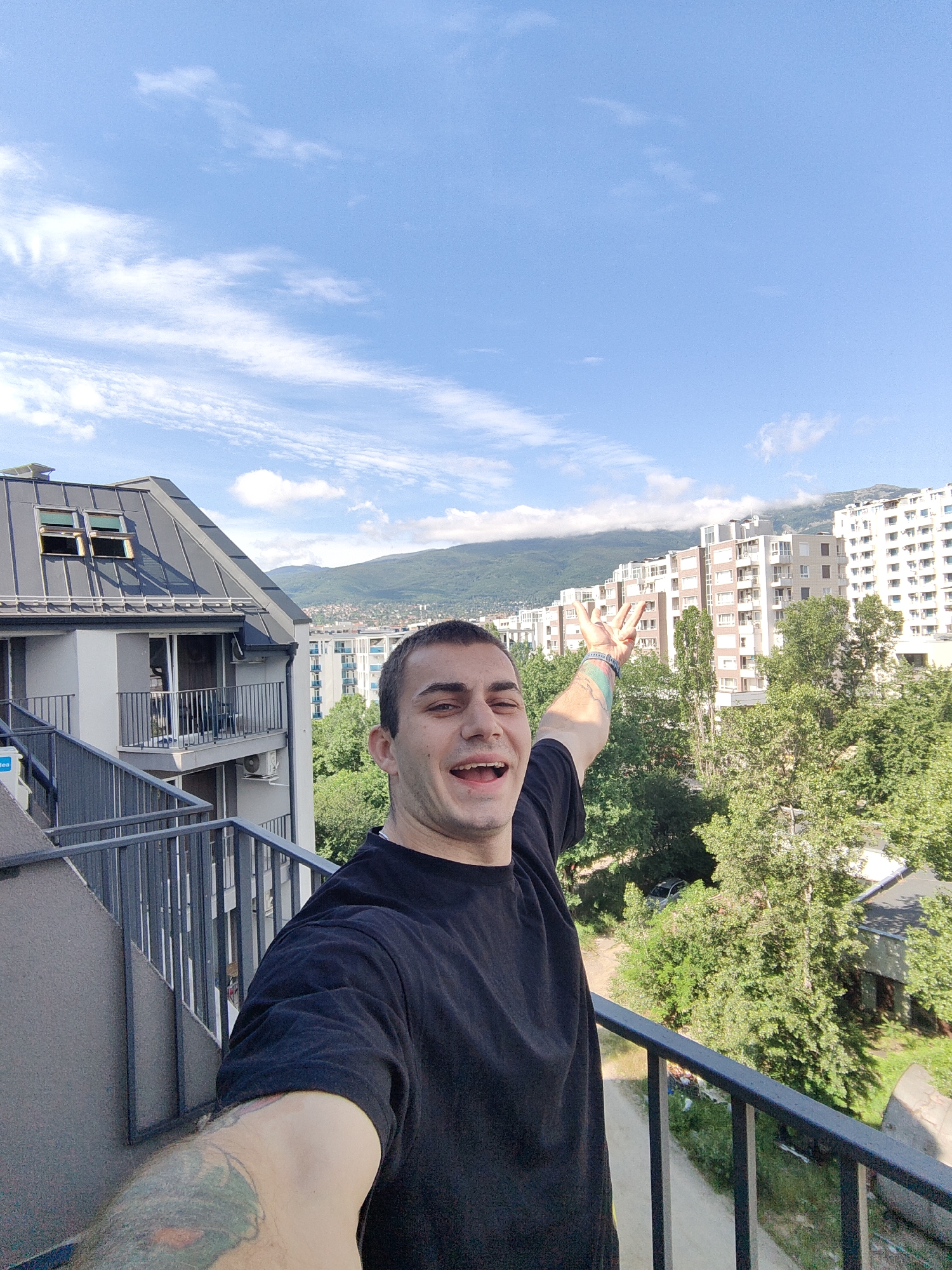 The image is a selfie taken from a balcony in a residential area of Sofia, Bulgaria. A man with short dark hair and a black t-shirt is the central subject, smiling and raising his right arm with his hand open, showing four fingers. He has tattoos on his left arm. The background features a modern apartment building with a grey, sloped roof and multiple windows, and further in the distance, several taller apartment buildings with light-colored facades and numerous windows. Lush green trees are visible between the buildings, and a mountain range with some cloud cover forms the backdrop. The sky is blue with scattered white clouds. A metal railing is present in the foreground, indicating the balcony from which the photo is taken.