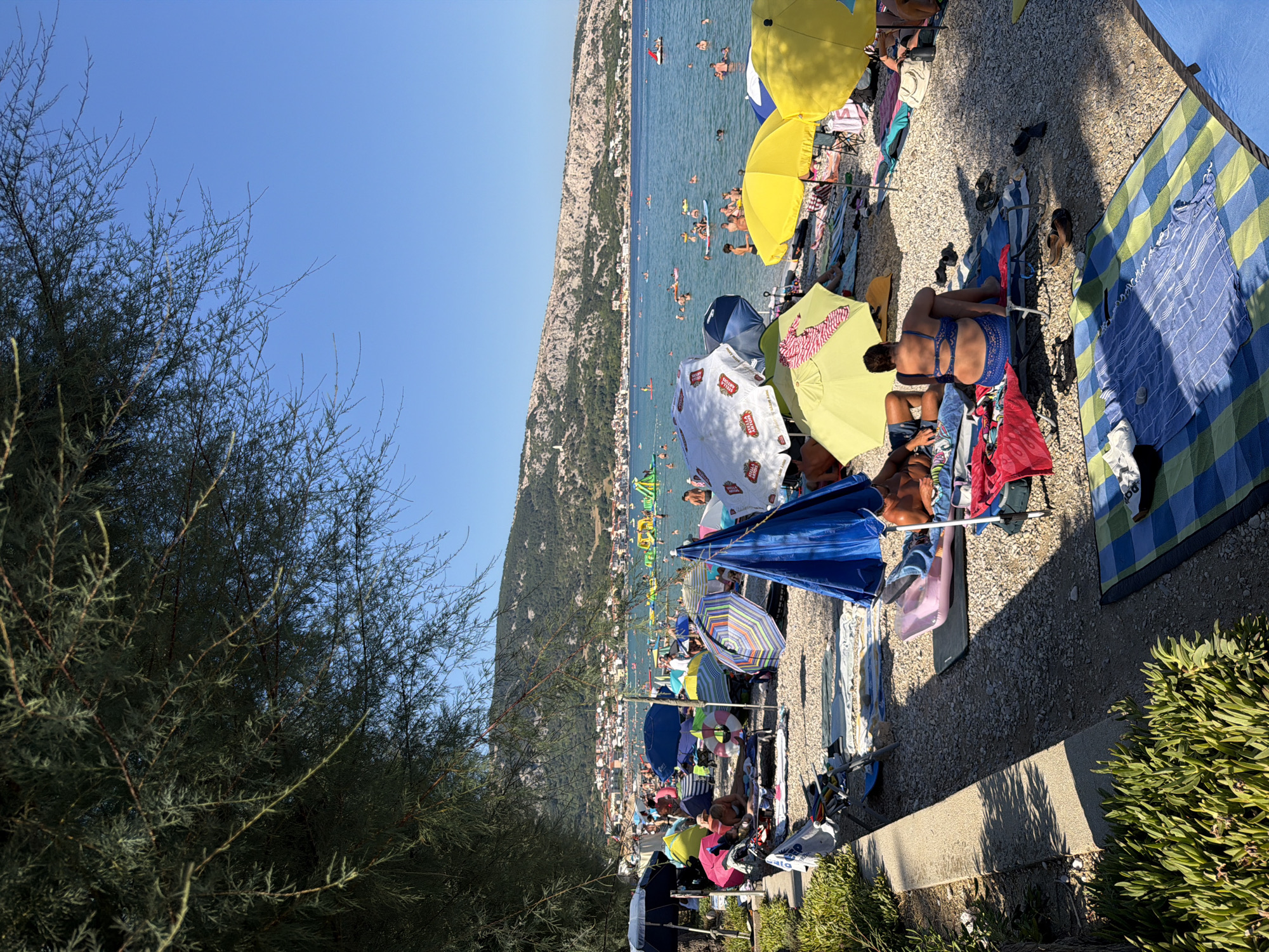 The image depicts a busy beach scene in Općina Baška, Croatia, on a sunny day. The foreground is dominated by a gravel beach lined with various beach umbrellas and mats. People are seen relaxing on lounge chairs and towels under the shade of the umbrellas. In the middle ground, the turquoise sea is filled with numerous swimmers and people enjoying various water activities, including paddleboarding and a floating water park. The background features a picturesque view of a town nestled against a hillside, with more buildings and lush greenery extending up the slopes. A clear blue sky stretches above the entire scene.