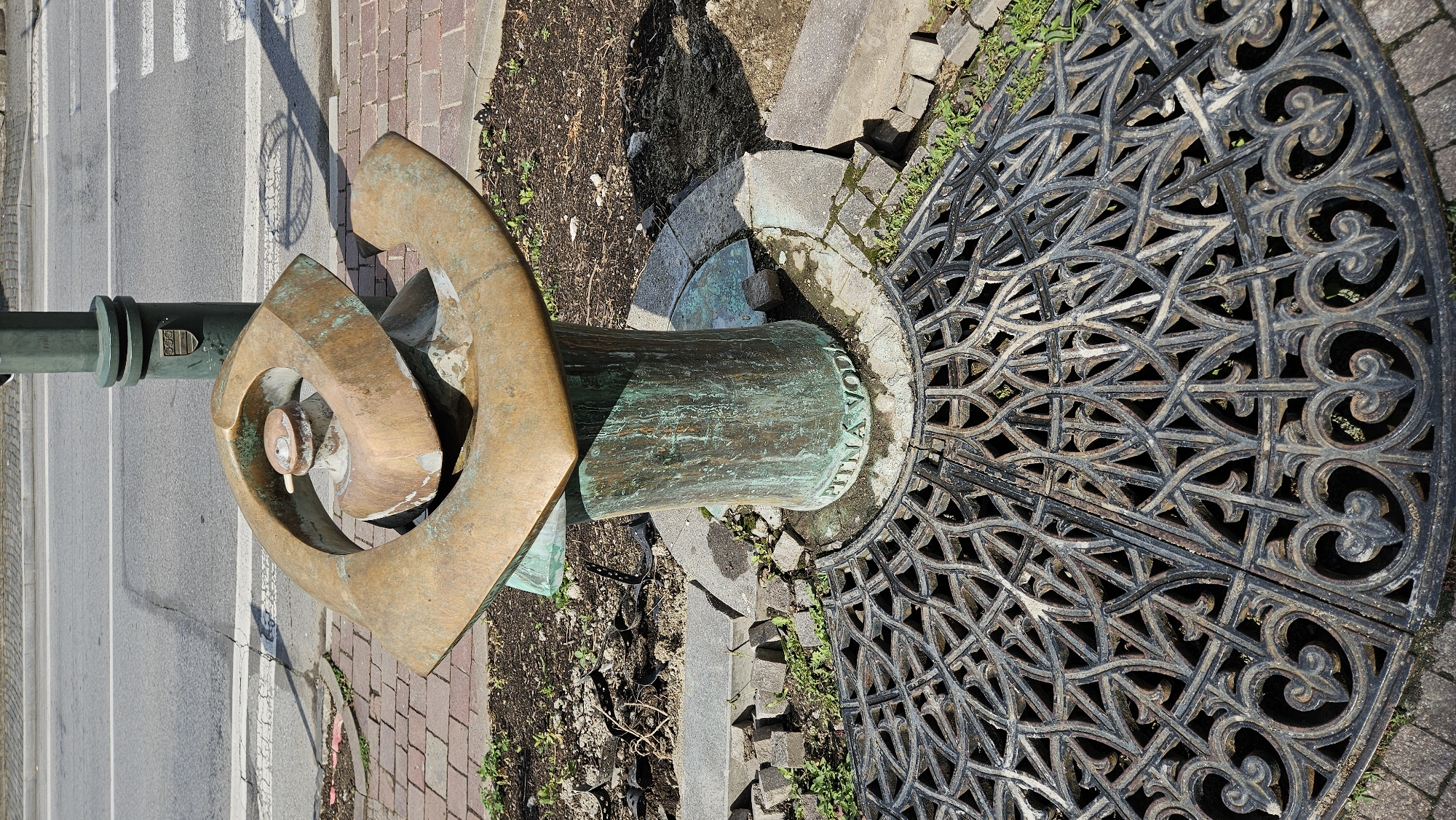 The image shows a public drinking fountain in the city of Prešov, Slovakia. The fountain is made of bronze and has a modern, abstract design, with a large, curved element surrounding a central spout. The base of the fountain is cylindrical and also made of bronze, showing signs of verdigris. Around the base, there is an inscription that reads "PITNÁ VODA", which translates to "drinking water". The fountain is situated on a circular paved area, surrounded by brickwork and bare earth, with some small plants growing in the soil. In the foreground, there is an ornate, black cast-iron grate covering a drain. Behind the fountain, a dark green lamppost stands tall, with a coat of arms mounted on it. Beyond the fountain and lamppost, a paved sidewalk and a road are visible, suggesting an urban setting. The lighting indicates it is daytime, with shadows cast by the fountain and lamppost. There are no people visible in the image.