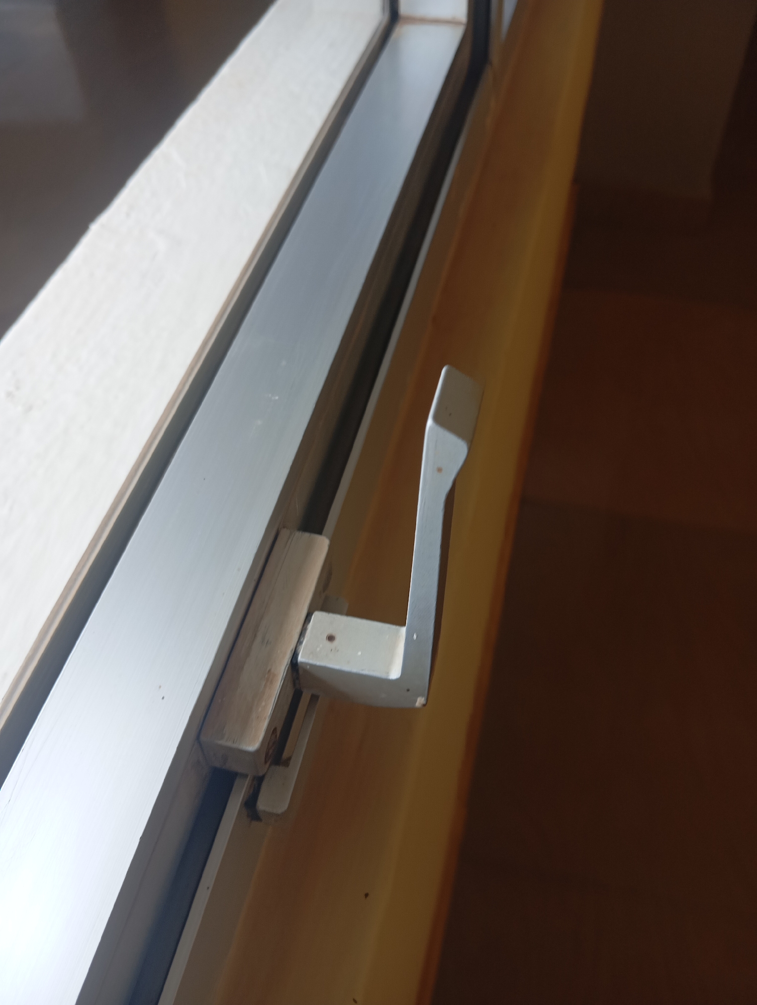 The image shows a close-up view of a window latch mechanism. The latch is made of metal and appears to be L-shaped, with one part extending horizontally and the other vertically. It is attached to a wooden frame which is part of the window. The window itself is framed by a light-colored material on the left and a darker, warm-toned wall or surface on the right. The overall scene suggests an interior view of a building, likely a room. There are no people visible in the image. The lighting is natural, coming from the window. The image does not provide any specific information that would allow it to be located in Jos, Nigeria.