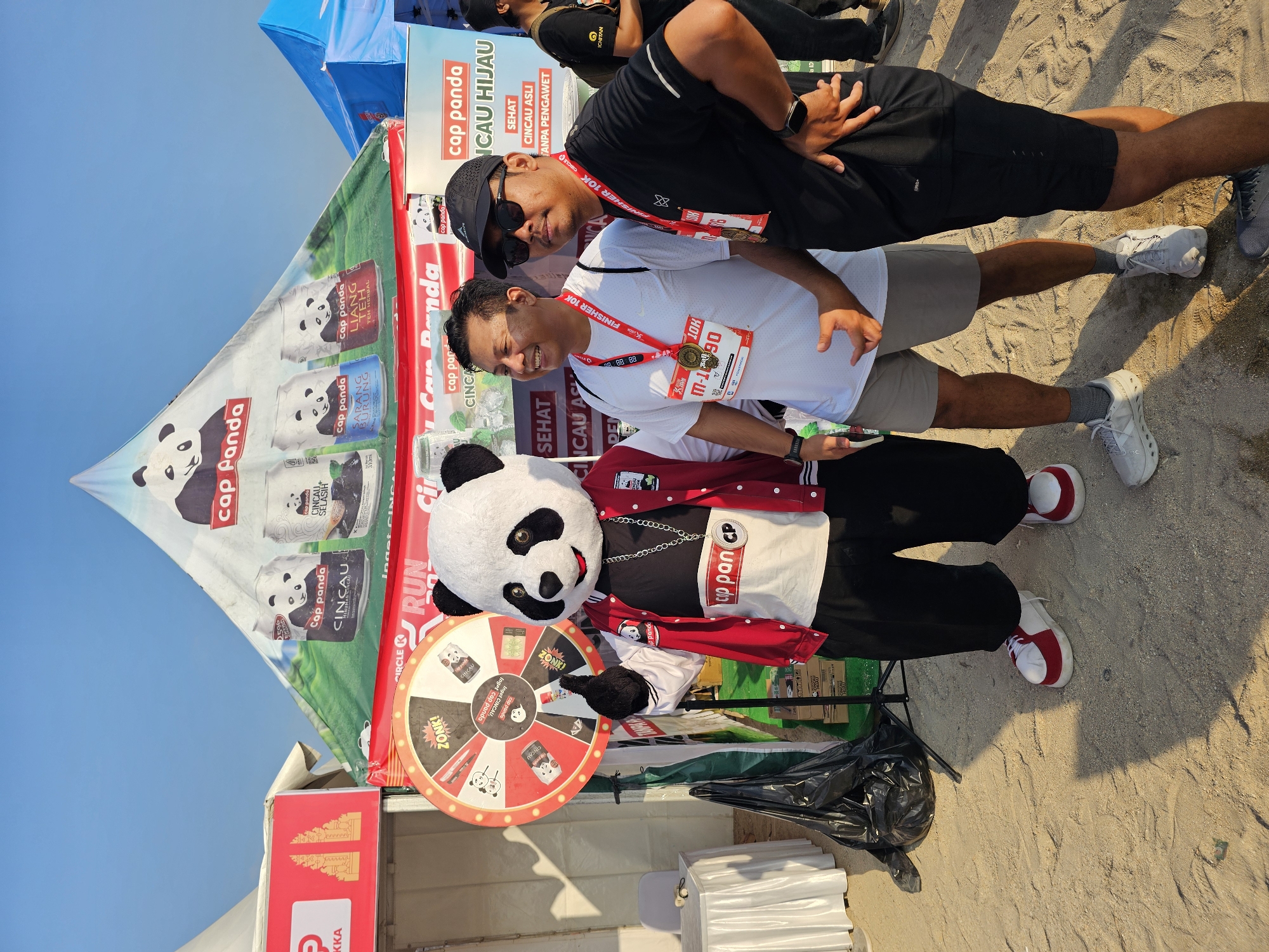 In Denpasar, Indonesia, at what appears to be an outdoor event with sandy ground, two men are posing for a photo alongside a person in a panda mascot costume. The panda mascot is wearing a red and white jacket with the "Cap Panda" logo, a white shirt with "Cap Panda" branding, and black pants with red and white shoes. One of the men, on the left, is wearing a white t-shirt with a medal around his neck, displaying "FINISHER 10K" and "M-1190." He has a medal pinned to his shirt. The man on the right is wearing a black t-shirt, sunglasses, a black baseball cap, and a medal around his neck, also showing "FINISHER 10K." A prize wheel with "ZONK!" and "Cap Panda" branding is visible to the left of the panda mascot. Behind them, a large tent with "Cap Panda" branding displays images of canned drinks. The sky is clear and blue.