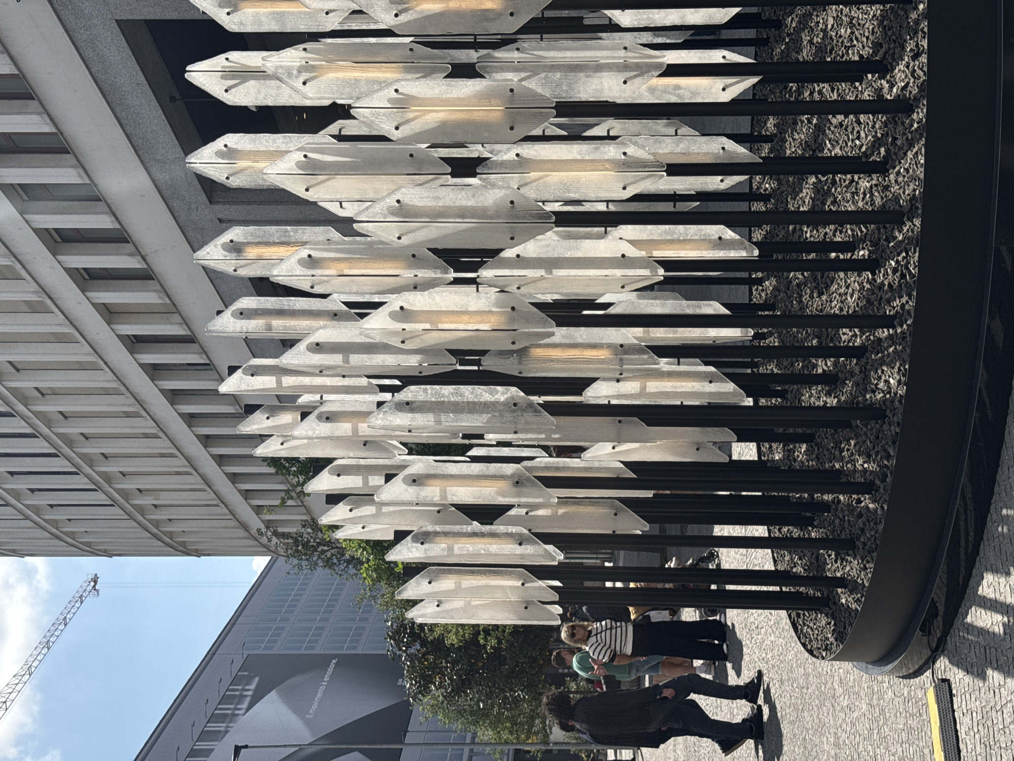 The image, originally rotated, depicts a contemporary urban public space, likely a plaza or walkway, under bright daylight. The dominant feature is a large, modern art installation or architectural element comprising numerous translucent panels. These panels, shaped like elongated rhomboids or flattened hexagons, are arranged in multiple horizontal rows and supported by dark vertical and horizontal rods, creating a structured, grid-like appearance. Each panel appears to house an internal light source, casting a soft, warm glow. The base of this installation is filled with dark gravel.
Several people are present in the scene, casually standing and walking around the installation. In the foreground, one person is seen from behind, wearing a black jacket and dark pants. Further back, another individual wears a striped top and sunglasses.
The background reveals modern buildings under a clear blue sky with some clouds. A tall, red and white construction crane is visible on the left side, indicating ongoing development or construction nearby. One of the buildings in the background has a large, grey facade with visible white text that reads "Il movimento è emozione," which translates from Italian to "Movement is emotion." This text, combined with the modern architecture and artistic installation, aligns with the provided location context of Milan, Italy. Sunlight casts clear shadows, suggesting a sunny day.