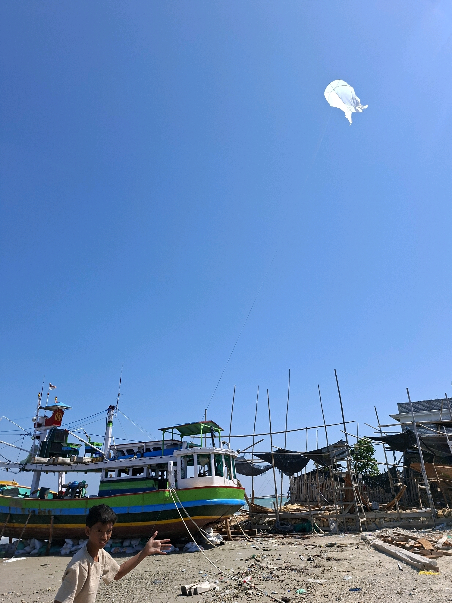 A young boy with dark hair, wearing a light-colored short-sleeved shirt, stands on a sandy beach in Camplong, Indonesia, with his left arm extended towards the viewer. Behind him, a large, multi-colored fishing boat is dry-docked, its hull painted in stripes of green, blue, and yellow. The boat is supported by wooden beams and white bags. To the right of the boat, an area under construction is visible, with wooden scaffolding and dark netting covering structures. The sky is clear and blue, with a single white kite flying high above. The beach is littered with debris, including wood, plastic, and rocks. The image appears to have been taken on a sunny day.