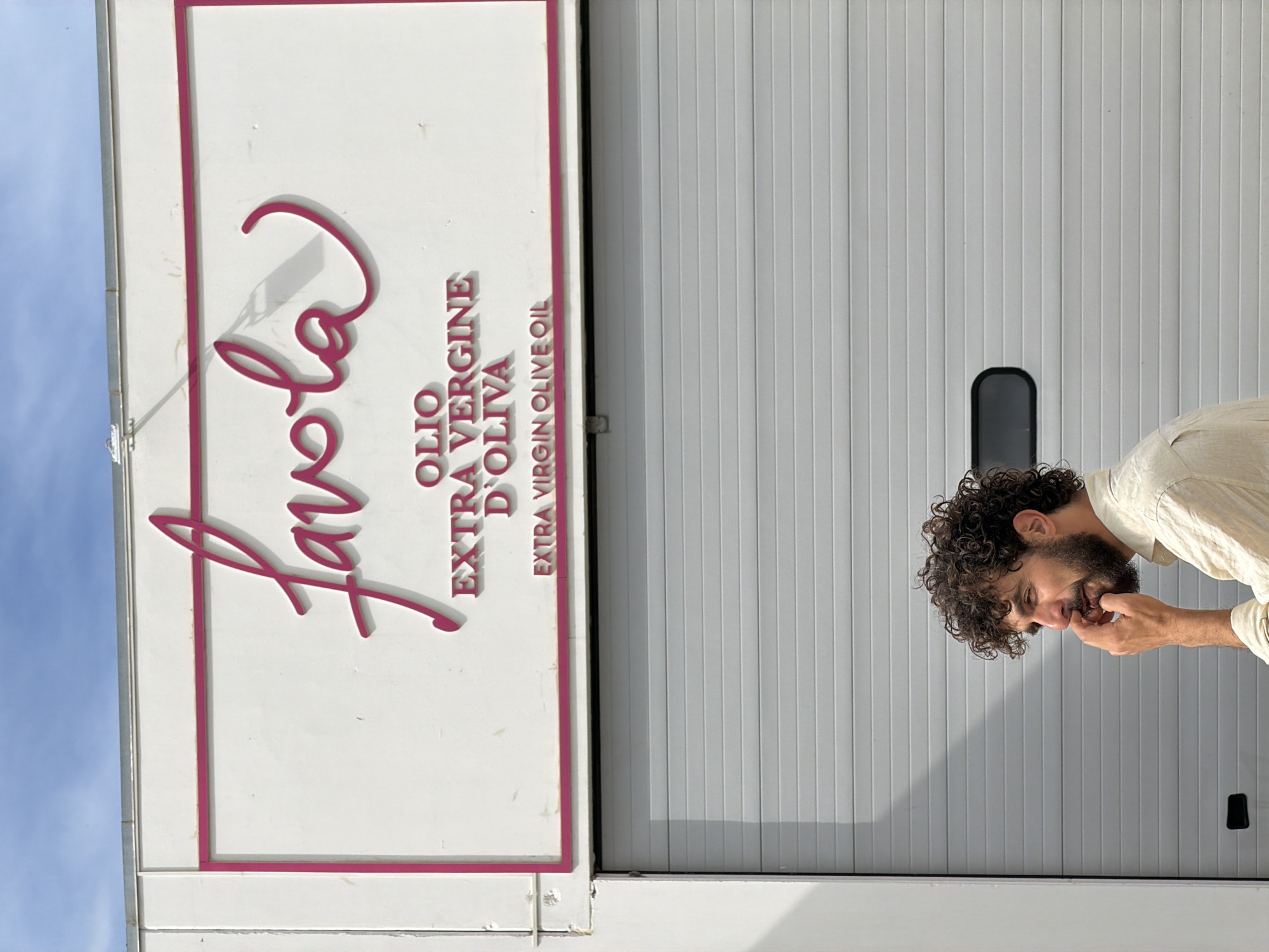 A man with dark, curly hair and a beard is partially visible on the right side of the frame, positioned in front of a light grey, vertically paneled wall or roll-up door. He is wearing a light-colored, possibly linen, long-sleeved shirt. The man is looking downwards, with his right hand near his chin and mouth, appearing pensive or contemplative.
To the left of the man and occupying a significant portion of the image, there is a large white rectangular sign with text in a vibrant pink color. The main word, "Favola," is written in a stylized script font. Below this, in capital letters, it reads "OLIO EXTRA VERGINE D'OLIVA" and underneath that, "EXTRA VIRGIN OLIVE OIL." The lettering has a slight shadow effect, giving it a three-dimensional appearance against the white background. The sign is bordered by a thin pink line.
The top left corner of the image shows a clear blue sky, indicating it is daytime. The overall setting appears to be outdoors, potentially at an olive oil facility in Solarino, Italy, as suggested by the sign's text.