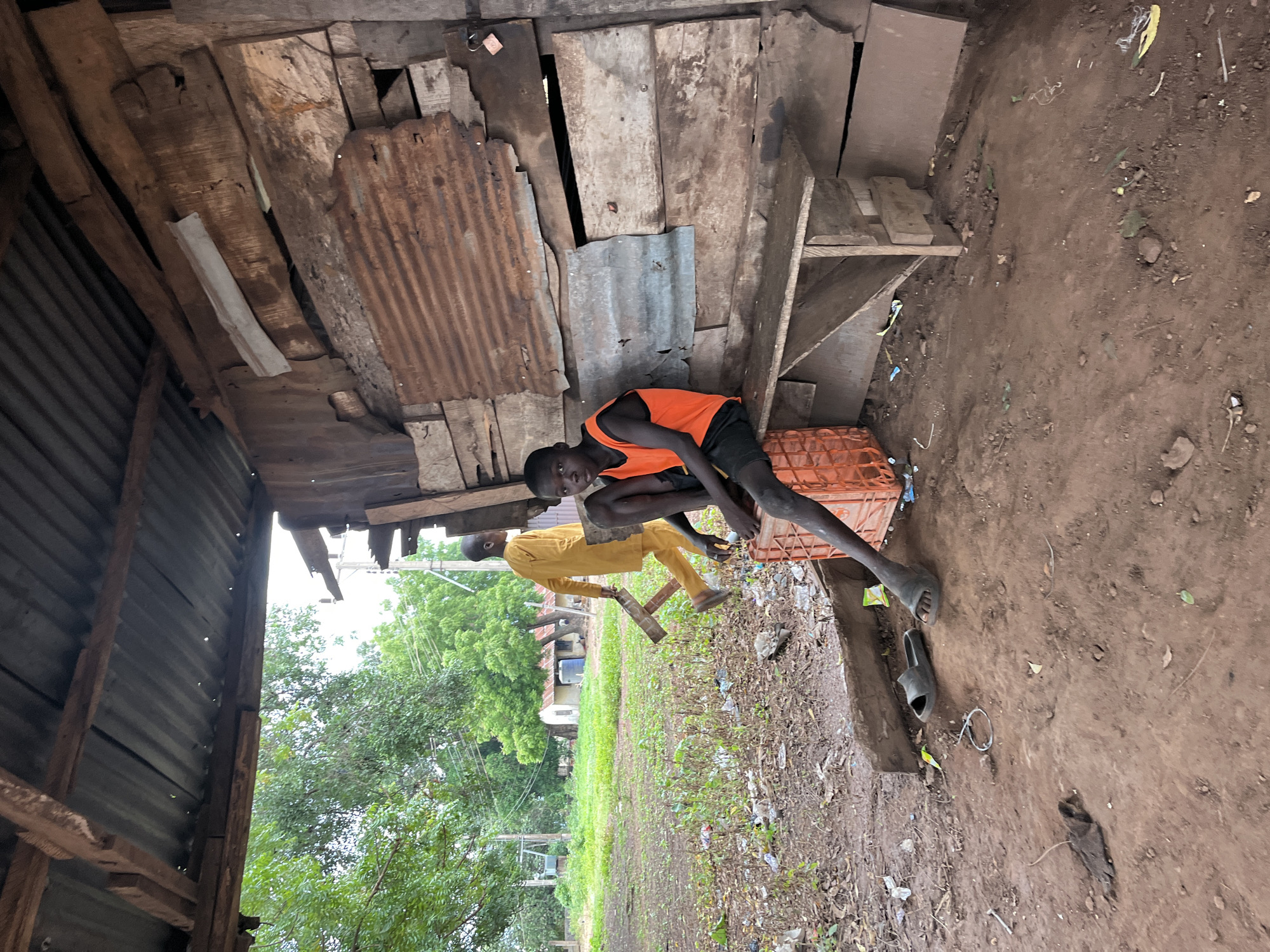 This image depicts an outdoor scene in Idah, Nigeria, featuring a makeshift structure and two males.
In the foreground and to the right, a rudimentary shelter is constructed from rough-hewn wooden planks and corrugated metal sheets. The roof, made of corrugated metal, is supported by visible wooden beams. The right wall of the structure is assembled from various weathered wooden boards and rusted corrugated metal panels. A small, unpainted wooden bench or shelf is affixed to this wall at a low height. A light-colored padlock is visible on a wooden plank in the upper right section of the wall.
A young male, appearing to be a child or adolescent, is seated on an orange plastic crate on the left side of the shelter. He wears an orange sleeveless top and dark shorts. His gaze is directed towards the viewer. His left leg is extended forward, and his right leg is bent. A pair of black slide sandals are on the reddish-brown earthen ground beside his feet; one is off his foot, and the other is partially on his right foot.
In the mid-ground, another male walks from left to right. He is dressed in a loose-fitting, long-sleeved, light yellow or mustard-colored garment. He carries a dark, angular wooden object, possibly a small frame or stool, in his right hand. His face is turned away from the camera.
The ground throughout the scene is composed of reddish-brown soil, interspersed with sparse patches of green vegetation and scattered debris, including plastic wrappers and discarded items. In the mid-ground and background to the left, an open, unkempt grassy area with numerous green-leafed trees is visible. Utility poles and overhead lines are present in the background. Further back, several simple structures with corrugated metal roofs are discernible, with one building featuring a blue tank or container beside it.
