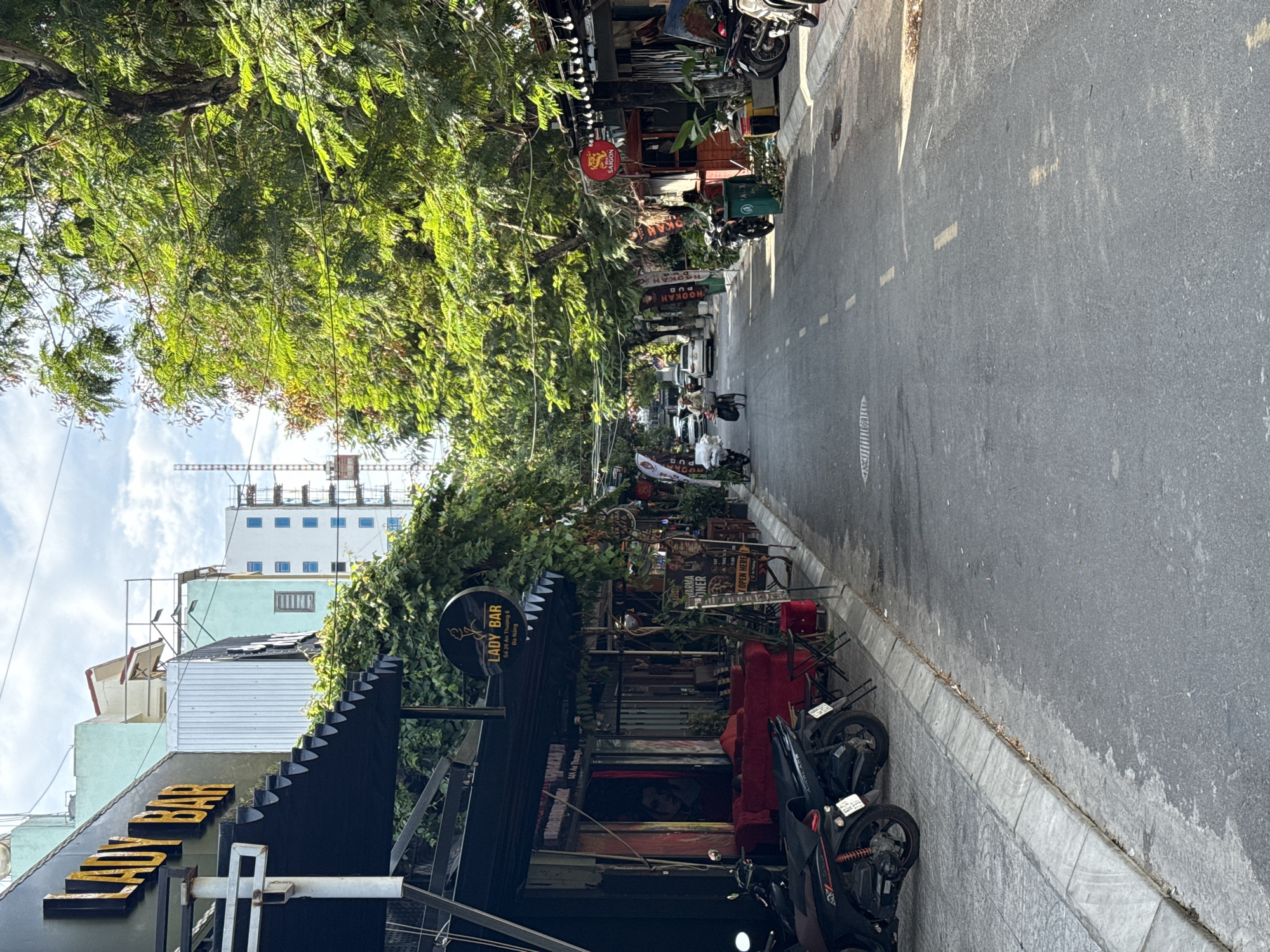 The image shows a street scene in Da Nang, Vietnam. On the left side of the street, there is a building with a sign that reads "LADY BAR". The bar has a black awning and red seating outside. Further down the street on the left, there are other businesses with signs and displays. On the right side of the street, there are more businesses, including one with a sign that says "SAIGON". There are trees lining the street, providing shade. Several motorbikes are parked along the side of the road. A few cars and motorcycles are visible in the street, and a person is riding a bicycle. The street is paved, and there are white and yellow lines painted on it to indicate lanes and pedestrian crossings. The sky is partly cloudy.