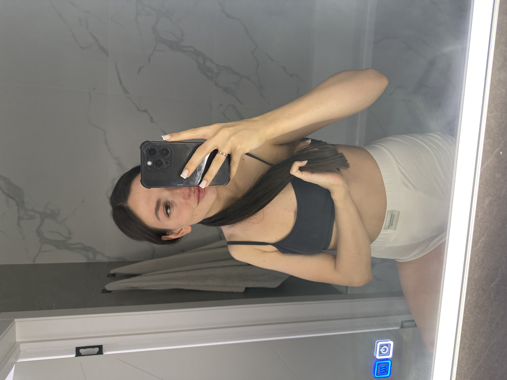 A young woman with dark hair pulled back in a ponytail is taking a mirror selfie in a modern bathroom. She is wearing a dark sports bra and white shorts. She is holding a smartphone in her right hand, which is angled to capture her reflection. Her left hand is holding her hair. The bathroom features marble-tiled walls, a white door with black hardware, and a lighted mirror with illuminated touch controls. The overall impression is one of a casual, private moment. There are no indications of the location being in the city of Estachi, Russia.