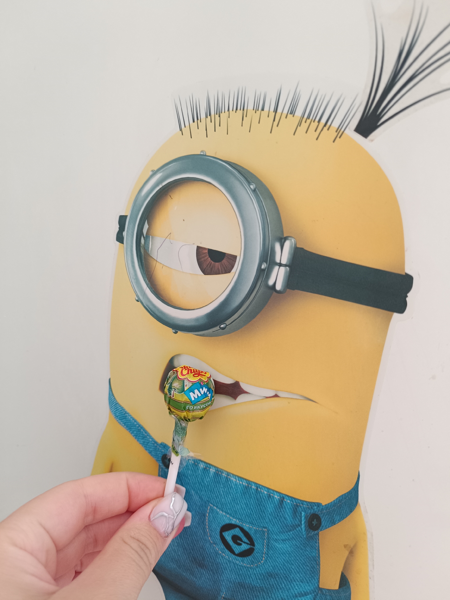 A photograph shows a cartoon Minion holding a lollipop. The Minion is yellow with one large eye behind a gray goggle. It has sparse black hair and is wearing blue overalls. The lollipop, which is a Chupa Chups brand, is being held up to the Minion's mouth by a hand with decorative fingernails. The packaging of the lollipop has Russian text, and the location is inferred to be Tobolsk, Russia.