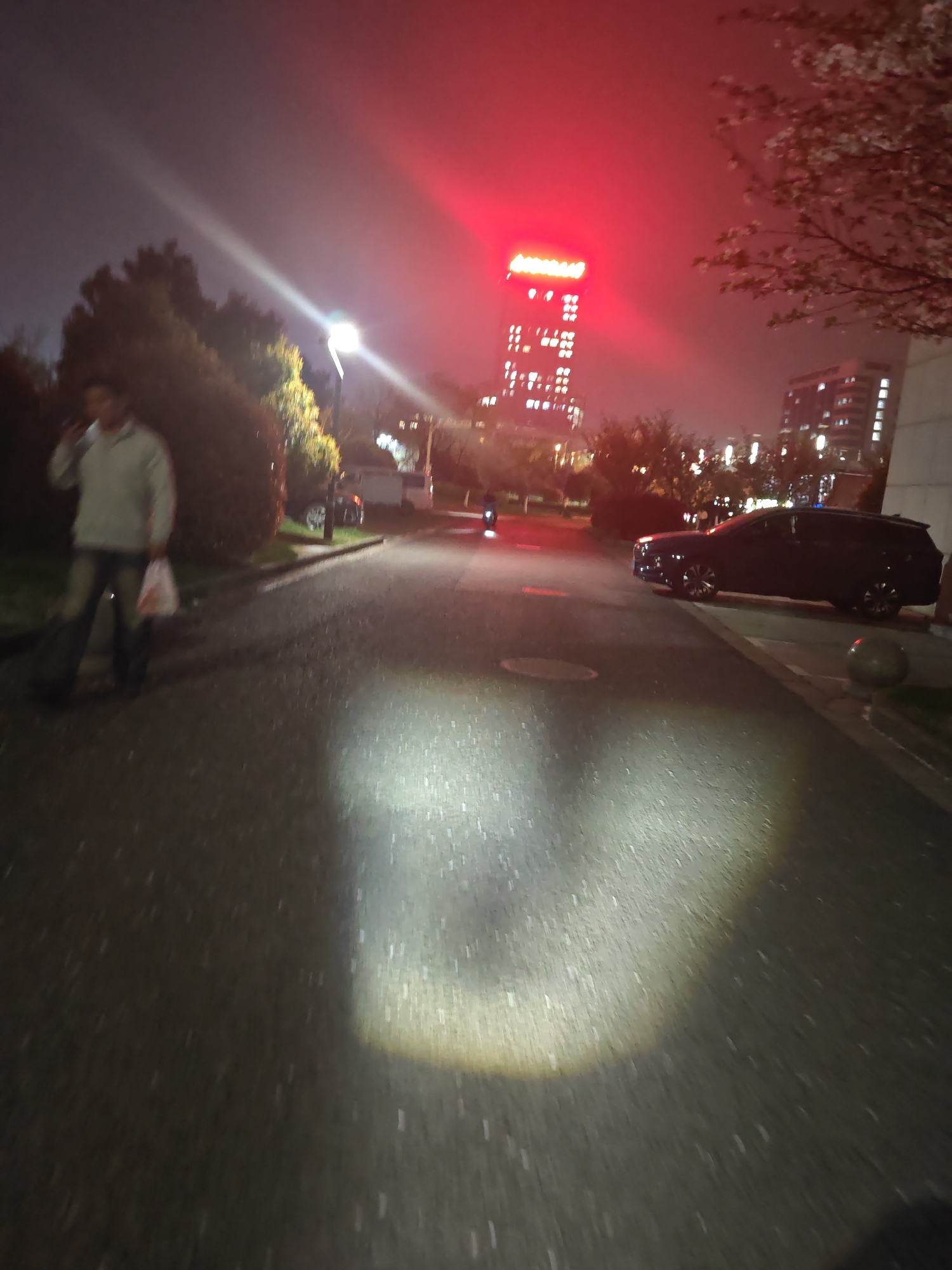 A nighttime scene in Jiangning District, China. A man, holding a white plastic bag, is walking and talking on a mobile phone on a paved road. A dark-colored station wagon is parked on the right side of the road, and a person is riding a motorcycle in the distance. A tall building with a red light on top stands in the background. The environment is dark with artificial lighting.