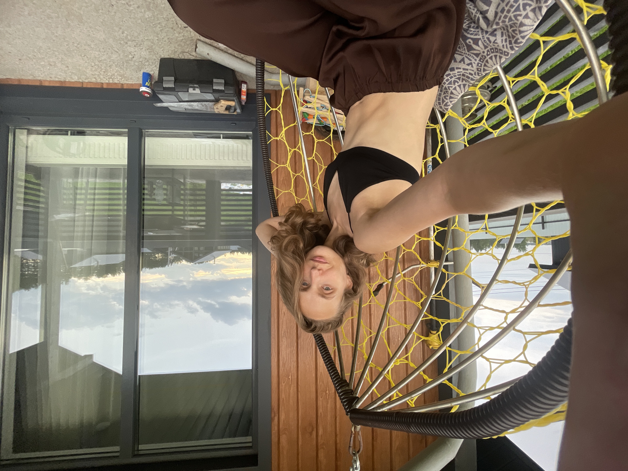 A young woman is captured in a selfie taken from a hanging chair on a covered porch. She is seated, wearing a black sports bra and loose-fitting brown pants. Her right arm is extended, holding the camera. The background reveals a wooden-paneled wall, a large window reflecting a cloudy sky and some houses, and a toolbox with a saw and can of spray paint on the ground. The hanging chair features a metal frame with yellow netting and a patterned cushion. The scene is outdoors, likely in a residential area, with a view of a suburban landscape.