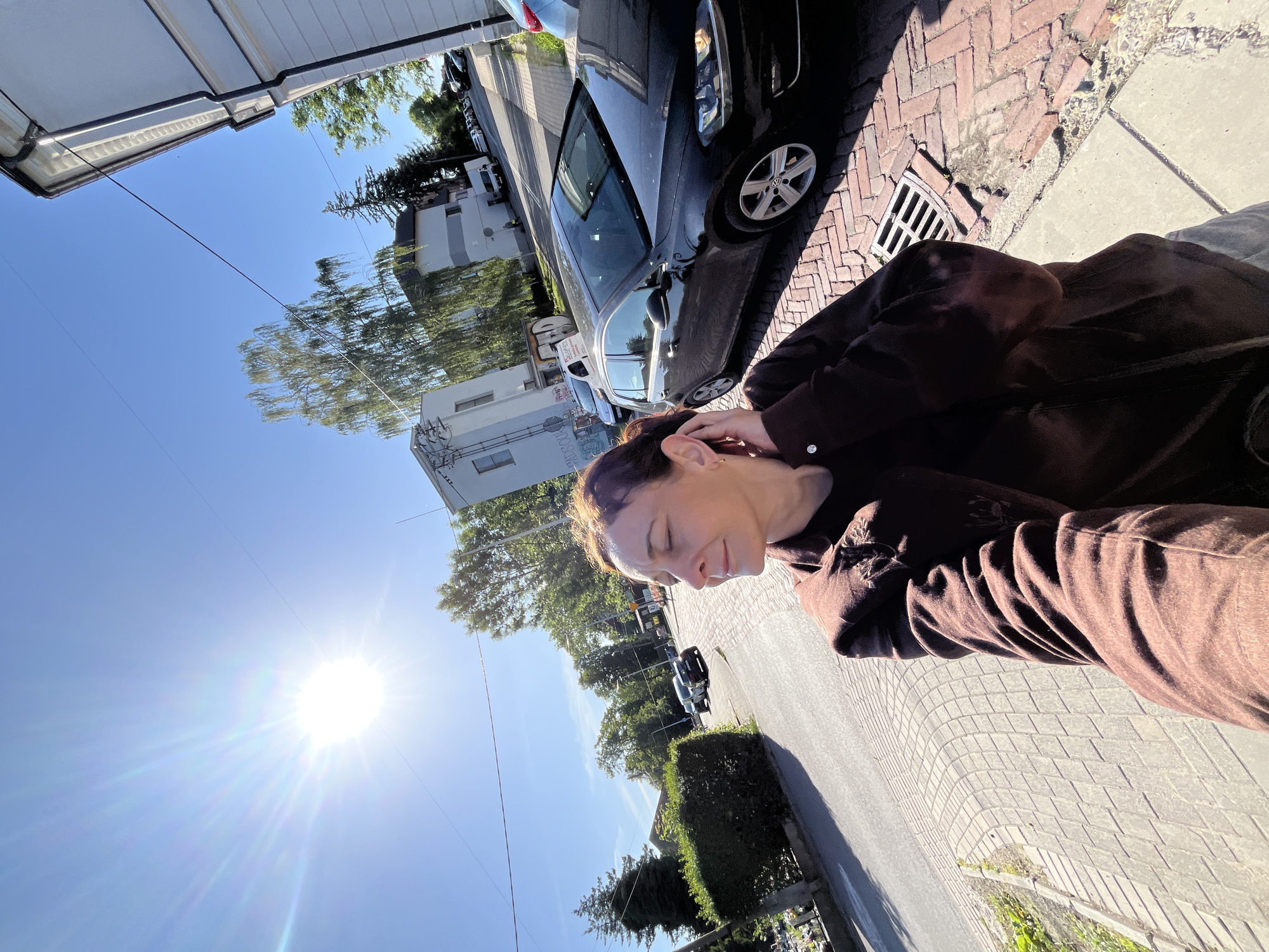 In a medium-shot, low-angle outdoor selfie, a woman with brown hair pulled back stands on a brick sidewalk. She wears a brown jacket and is reaching up to touch her ear, her eyes closed as if basking in the sun. The sun, a bright, starburst of light, dominates the upper left of the frame, casting long shadows. Behind her, a street curves to the left, lined with parked cars and buildings. A black Volkswagen Golf is parked on the right side of the frame, its headlights on. The background includes trees and a clear blue sky. The overall scene suggests a sunny day in a residential or commercial area.
