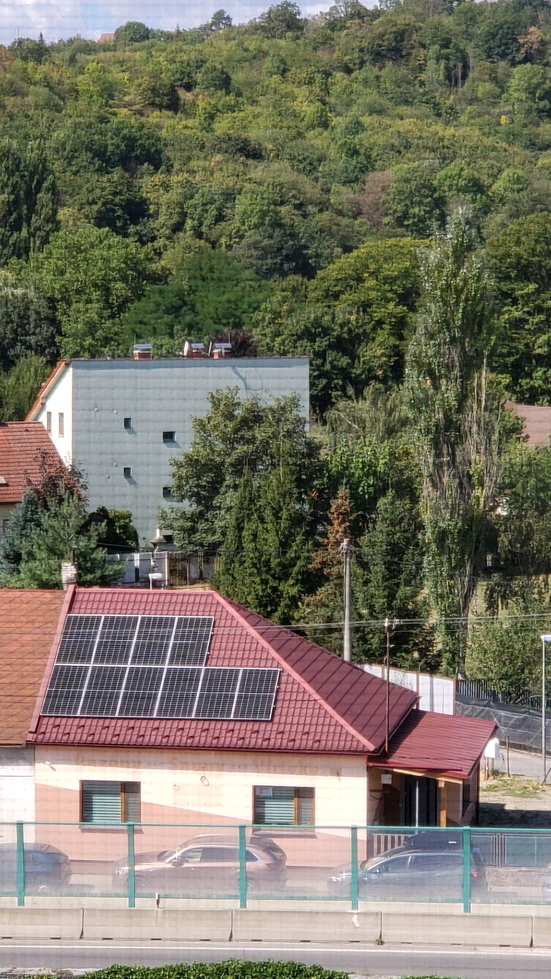 The image shows a residential building with a red tiled roof and solar panels. The building has a light-colored facade with two windows on the lower level. In front of the building, there is a green fence and several cars parked behind it. In the background, a dense green forest covers a hillside, with a taller, slender tree to the right of the frame. A larger, grey building with several small square windows is visible behind the foreground house and to the left. The sky is partly cloudy. This scene is located in the city of Prešov, Slovakia.