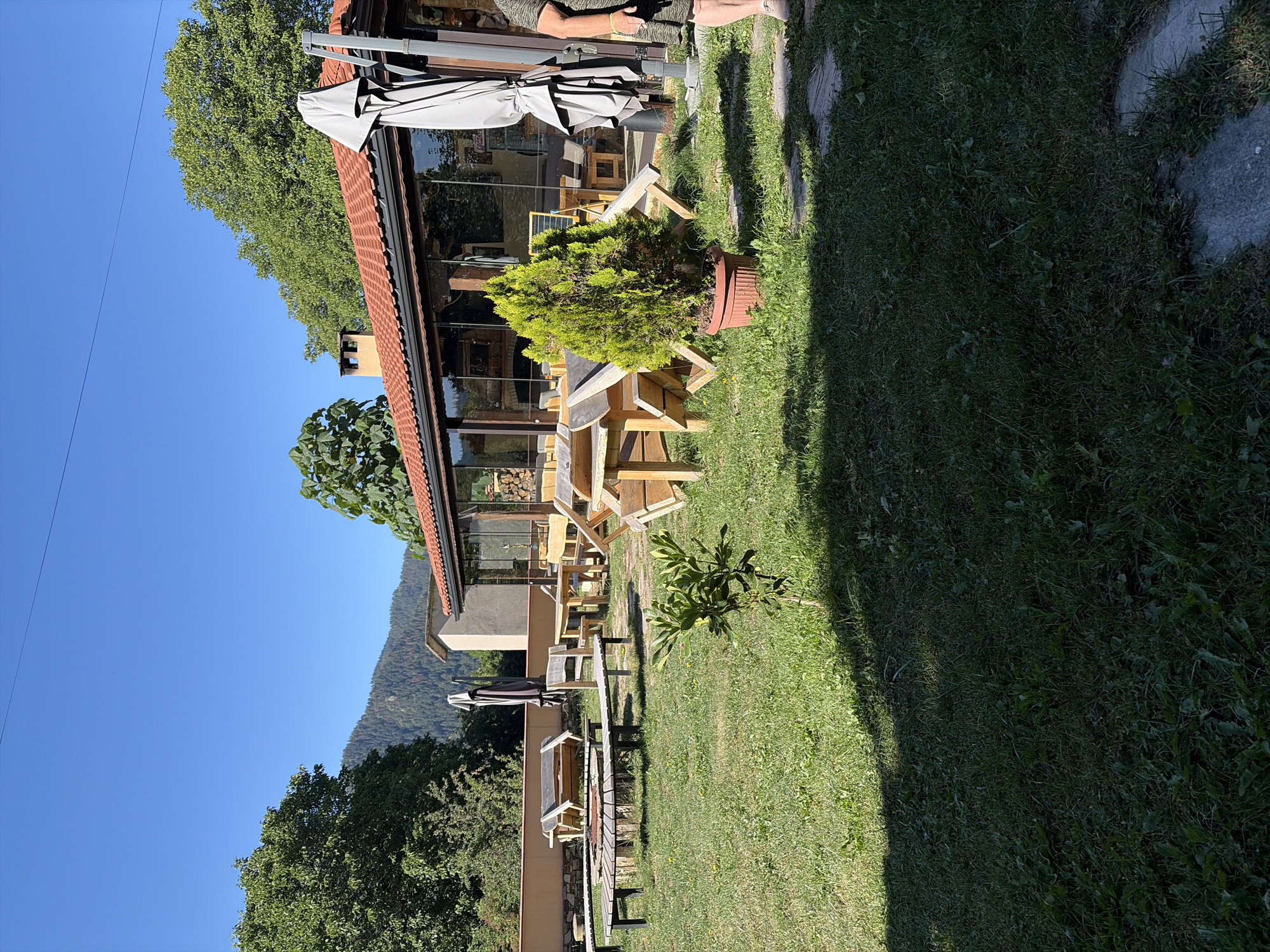 The image depicts an outdoor seating area of a restaurant or cafe in Smolyan, Bulgaria. The scene is set on a sunny day with a clear blue sky. In the foreground, a grassy area is sparsely populated with small plants and some stone pavers. Several rustic wooden benches and tables are arranged around a central fire pit made of logs.
In the mid-ground, a building with a red-tiled roof features a glass-enclosed section, likely housing the restaurant's interior dining area. A tall, potted evergreen tree stands prominently in front of the glass. Next to the building, a grey outdoor umbrella is folded and positioned upright. A person, partially visible on the right edge of the frame, is wearing a patterned dress and appears to be standing.
The background reveals a lush green landscape with mature trees and a distant, tree-covered mountain. The overall impression is of a relaxed, natural setting for outdoor dining.