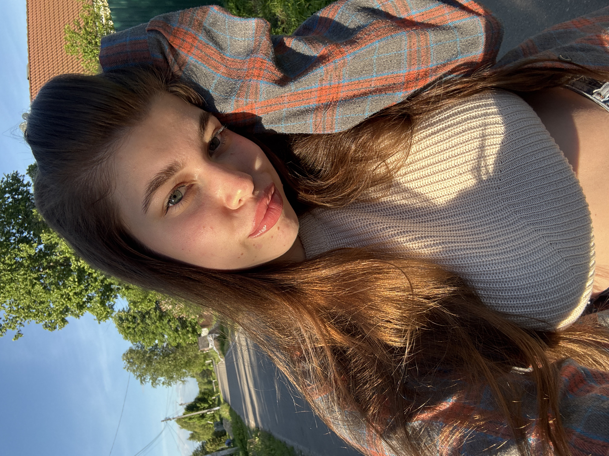 This vibrant outdoor portrait captures a young woman with long, warm brown hair and light-colored eyes, seemingly in a rural or suburban area of Мещёры, Russia. The image is oriented such that she is lying down, positioned with her head towards the upper left, gazing directly and softly at the camera. She has fair skin, with visible freckles, and her lips are slightly parted, conveying a relaxed and calm expression.
She is dressed in a light-colored, possibly beige or light grey, knitted sweater, worn beneath a plaid shirt patterned with shades of red, orange, blue, and grey. Her hair and the scene are illuminated by the warm, directional light of what appears to be the golden hour – either late afternoon or early morning – casting prominent shadows and giving her hair a radiant sheen.
The background features a clear, bright blue sky and lush green trees. A paved road stretches into the distance, bordered by a green fence. A glimpse of a building with a reddish-brown tiled roof and a utility pole with wires are also visible. The clear sky indicates sunny weather. There is no discernible text within the image.