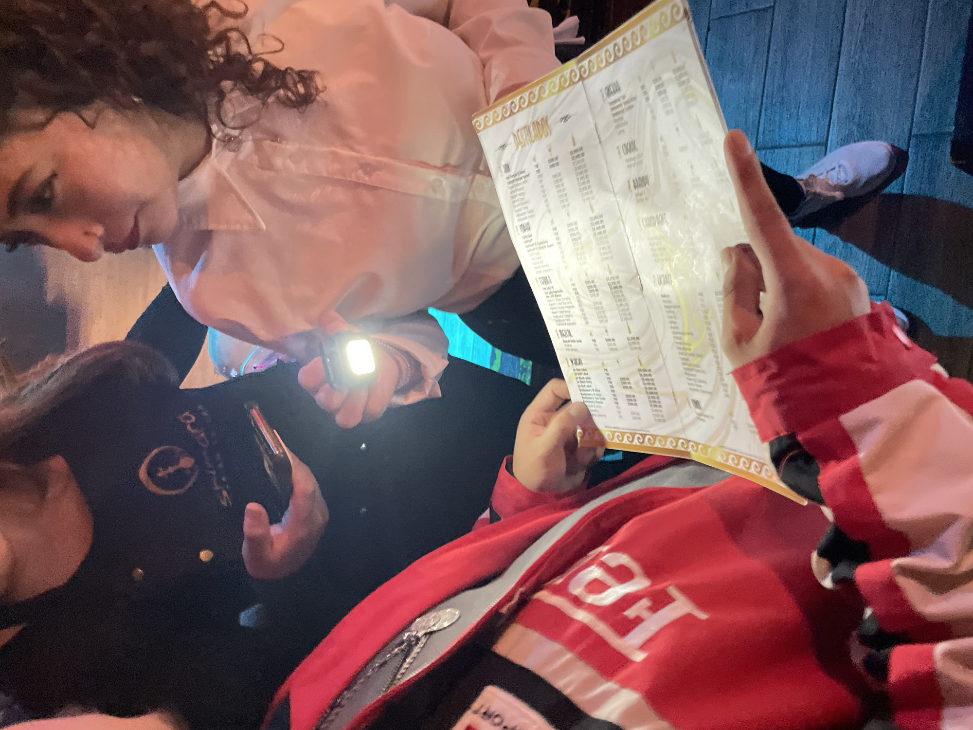 The image depicts three individuals in a dimly lit setting, possibly a restaurant in Mexico City, Mexico. A woman with curly hair, wearing a white blouse, holds a flashlight to illuminate a menu for a person in a red jacket who is pointing at the menu. A third person in a black shirt with a logo is holding a smartphone. The environment appears to be a dark interior with blue accents in the background.