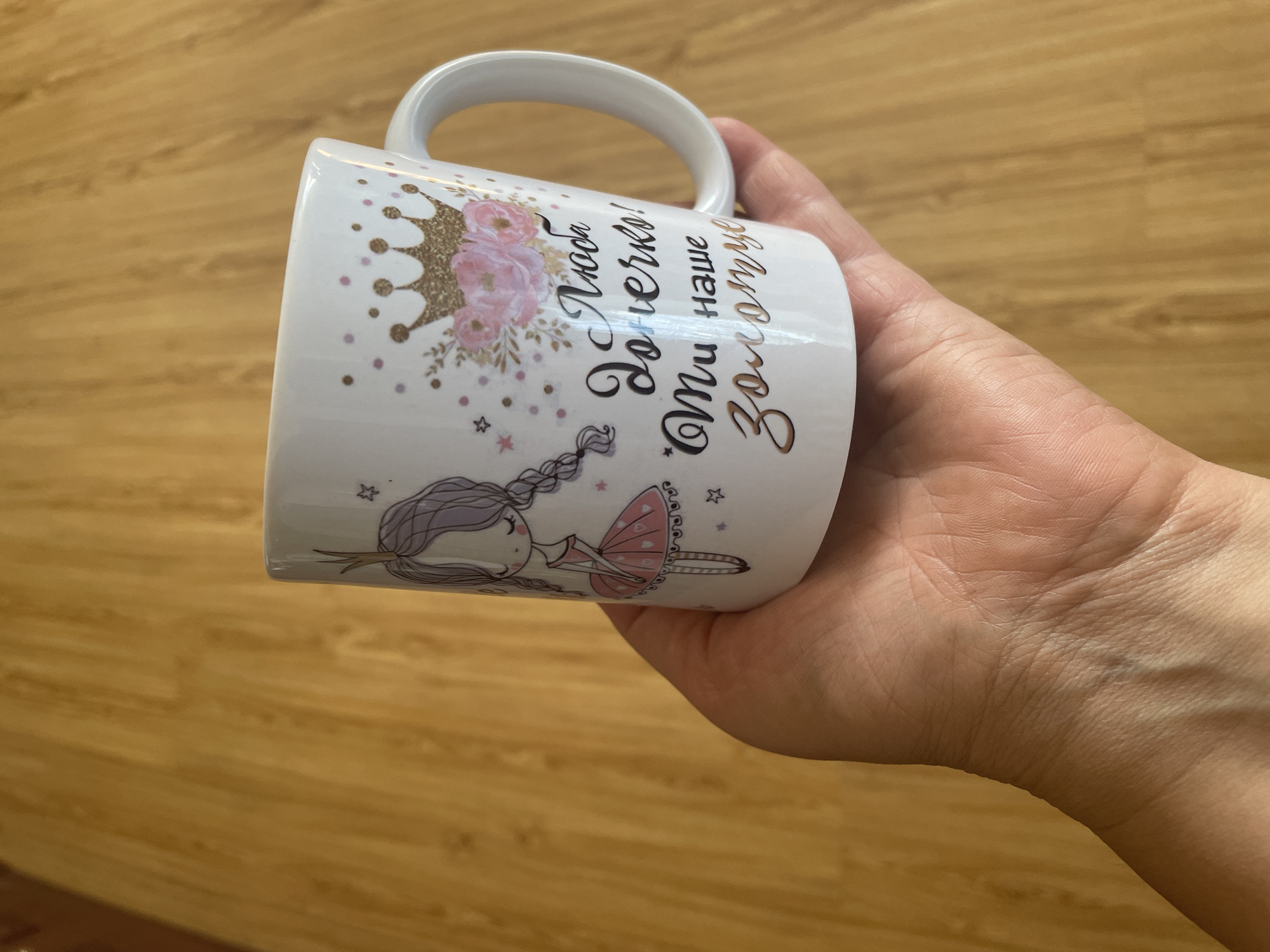 A hand holds a white ceramic mug with a drawing of a girl in a pink dress and a crown, and the words "Люба донечко! Ти наше золотує" in Russian. The background is a wooden surface. This image was captured in Shpykolosy, Ukraine.