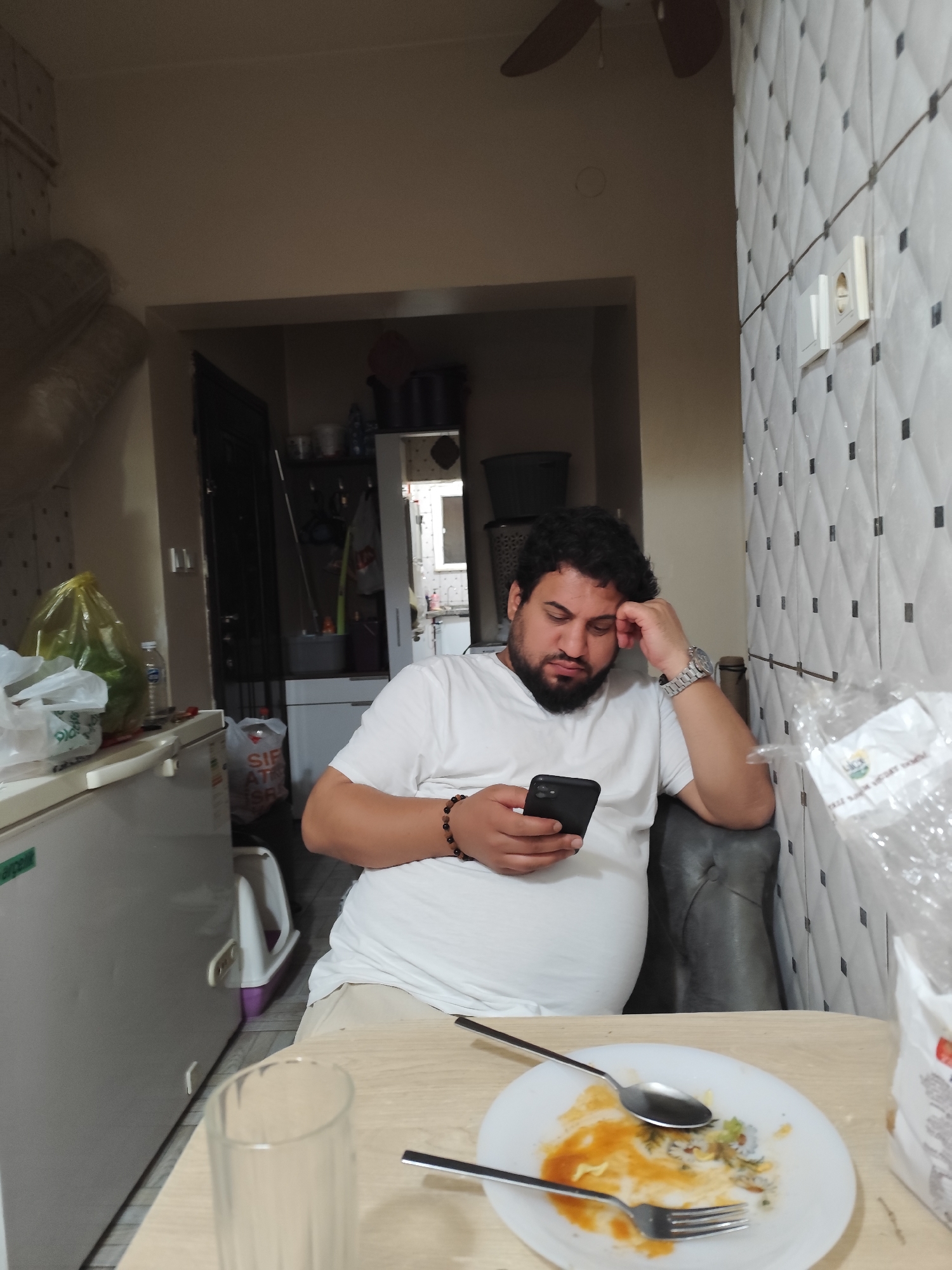A man with dark, curly hair and a beard, wearing a white t-shirt, is seated indoors, engrossed in his smartphone. He rests his head on his left hand, elbow supported by a dark grey armchair or sofa, conveying a contemplative or slightly tired expression as he focuses on his device. He wears a silver-toned watch on his left wrist and a beaded bracelet on his right.
The scene appears to be a casual dining or living area within a home in Sarıyer, Turkey. In the foreground, a light wooden table holds a white plate with remnants of a saucy meal, a spoon, and a fork, suggesting he has recently eaten or paused his meal. An empty clear glass is also on the table. To the left, a white refrigerator or deep freezer is visible, with a yellow plastic bag, another white plastic bag, and a water bottle on its top surface. A dark doorway in the background leads to another room, likely a kitchen or utility area, where shelves with various items are partially visible.
The wall to the right is covered with distinctive white diamond-patterned tiles interspersed with black square accents, and two electrical outlets are mounted on it. A brown ceiling fan is partially visible above. On the floor behind the refrigerator, a white plastic bag bears fragmented text, possibly "SIFIR ATIK BAŞLADI" (Zero Waste Started). Another white plastic bag with the word "LİCE" and other unreadable text is on the table to the right, partly out of frame. The image captures a quiet, everyday moment indoors, likely during the day.