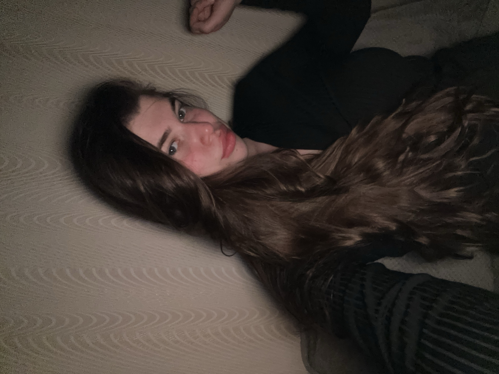 A young woman is lying on a cream-colored textured surface. She has long brown hair and is wearing a black long-sleeved shirt. Her right arm is bent at the elbow and raised slightly. The scene is dimly lit. The location is not apparent from the image.