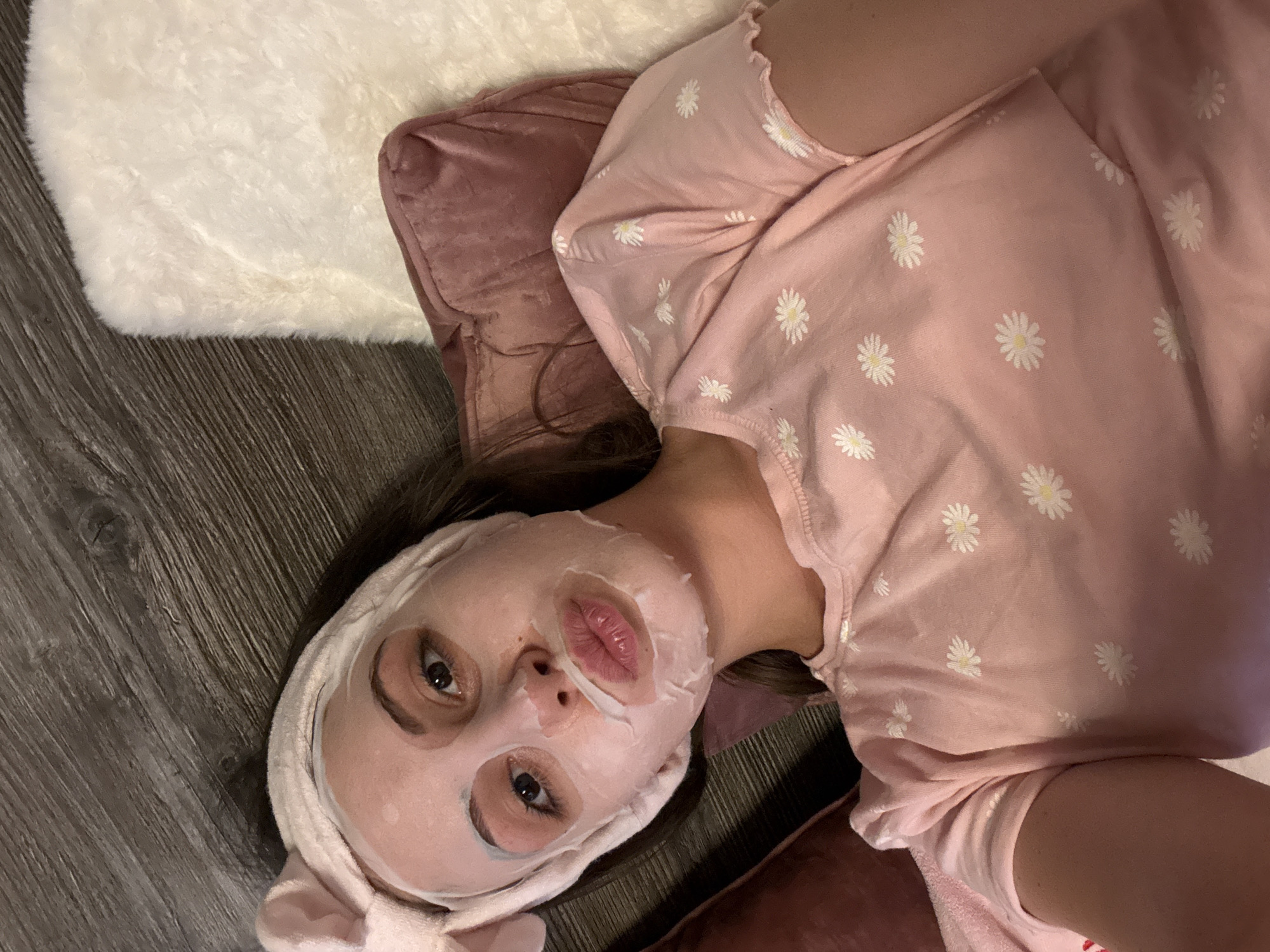 A young woman with a sheet mask and a pink headband with bunny ears is lying down on a wooden floor next to a plush white rug and a pink pillow. She is wearing a pink t-shirt with a daisy print. The scene appears to be indoors, possibly a bedroom or living room. There is no visible indication of the specific location in Bulgarovo, Bulgaria.