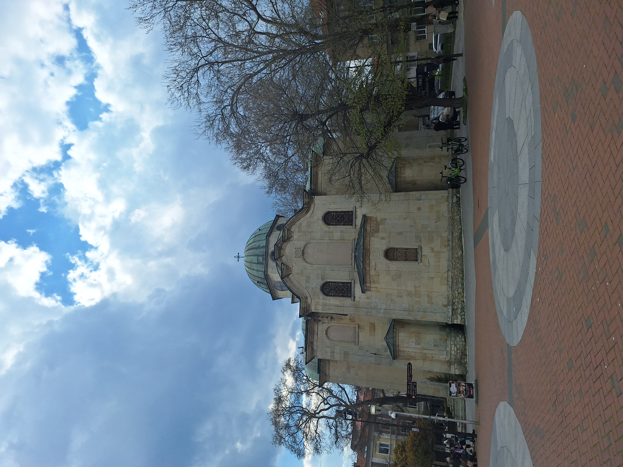 The image shows the Dormition of the Mother of God Cathedral in Varna, Bulgaria. The cathedral, constructed of light-colored stone, is seen from a low angle, with its green dome prominent against a sky filled with white and gray clouds. Bare-branched trees frame the cathedral against the sky.
A brick-paved public space is visible on the right side of the image, with circular patterns inlaid in gray stone. Bicycles are parked nearby. A street with cars and pedestrians is visible in the background to the right of the church. Street signs are visible on a pole, suggesting the presence of an urban environment.