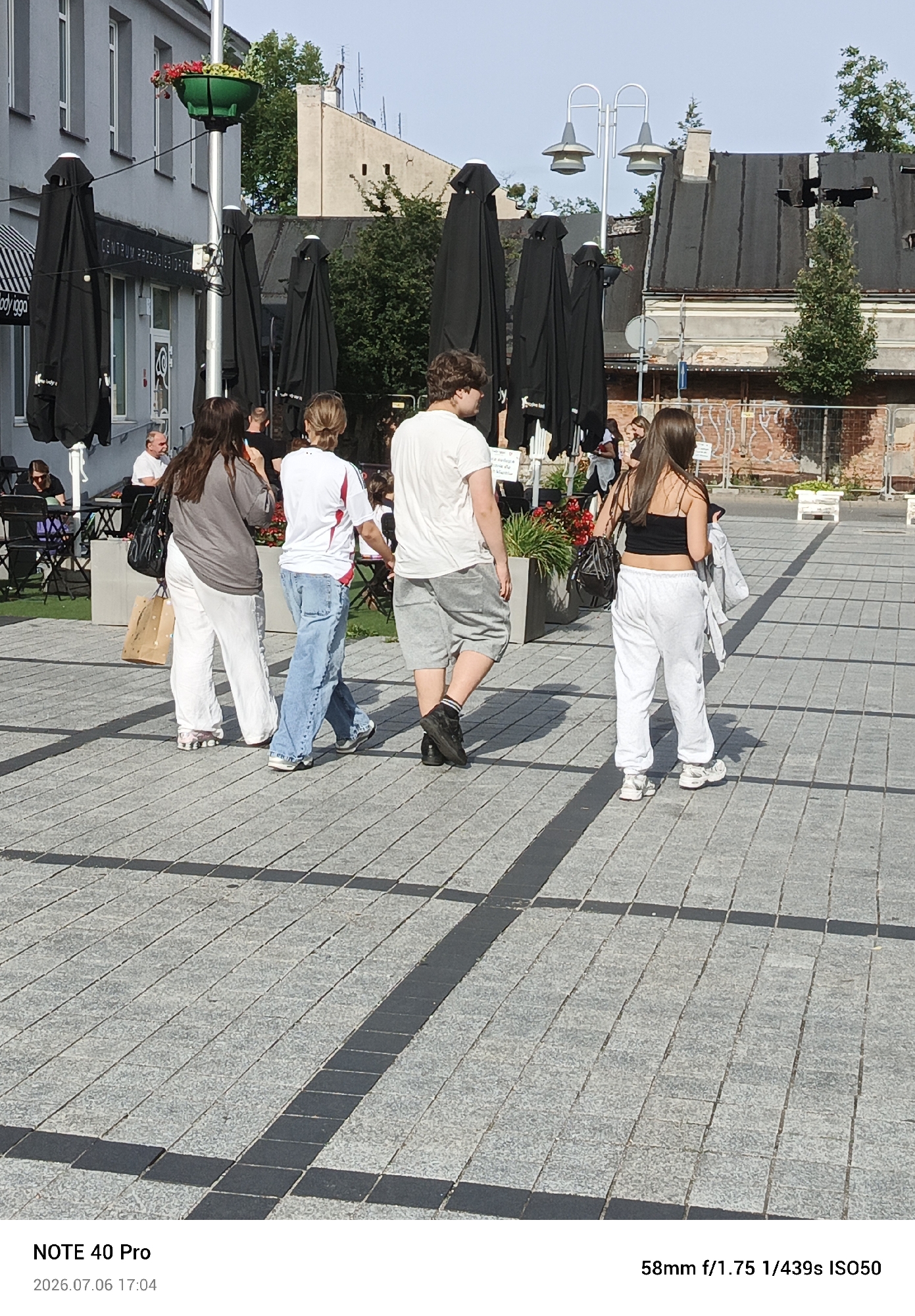 The image depicts a paved public square with several people walking and standing. In the foreground, a group of four young adults are visible. The person on the far left, seen from the back, wears a grey t-shirt and white trousers. Next to them, a person in a white t-shirt with red and black accents and blue jeans is walking away from the camera. To their right, a person in a white t-shirt and grey shorts walks towards the right of the frame. The person on the far right, also seen from the back, wears a black crop top and white trousers. They are holding a jacket in their left hand. In the background, a building with a grey facade and black umbrellas set up outside a cafe are visible. Further in the background, damaged buildings with dark roofs and exposed brickwork are present. The ground is paved with grey and black square tiles, forming a patterned surface. A lamppost with a green planter containing red flowers stands to the left of the frame. The scene appears to be an outdoor urban setting under a clear sky, likely during daylight hours. The text overlay at the bottom indicates "NOTE 40 Pro 2026.07.06 17:04" and "58mm f/1.75 1/439s ISO50". This image was taken in Piaseczno, Poland.