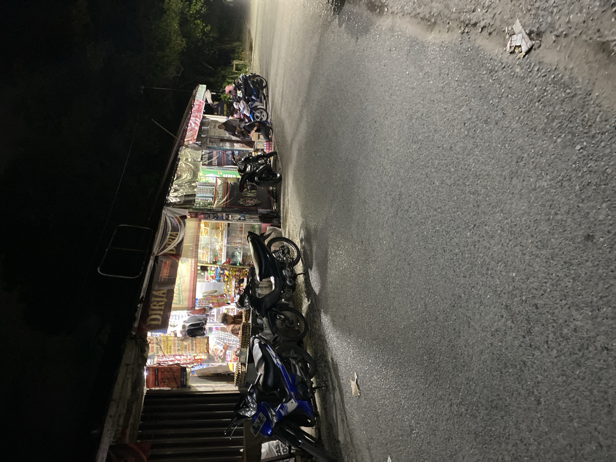 A night-time, eye-level, full-shot scene depicts a roadside store with several motorcycles parked in front of it. The store, partially illuminated by its internal lighting, displays various goods on shelves behind a counter. A few people are visible near the counter and seated on the ground, interacting with each other. A blue and black motorcycle is parked on the left, followed by a black motorcycle. Further down the road, two more motorcycles, one blue and one black, are parked, and two individuals are seated on the ground near them. The scene is set on a paved road with a slight incline and scattered debris. Trees are visible in the background, and the sky is dark.