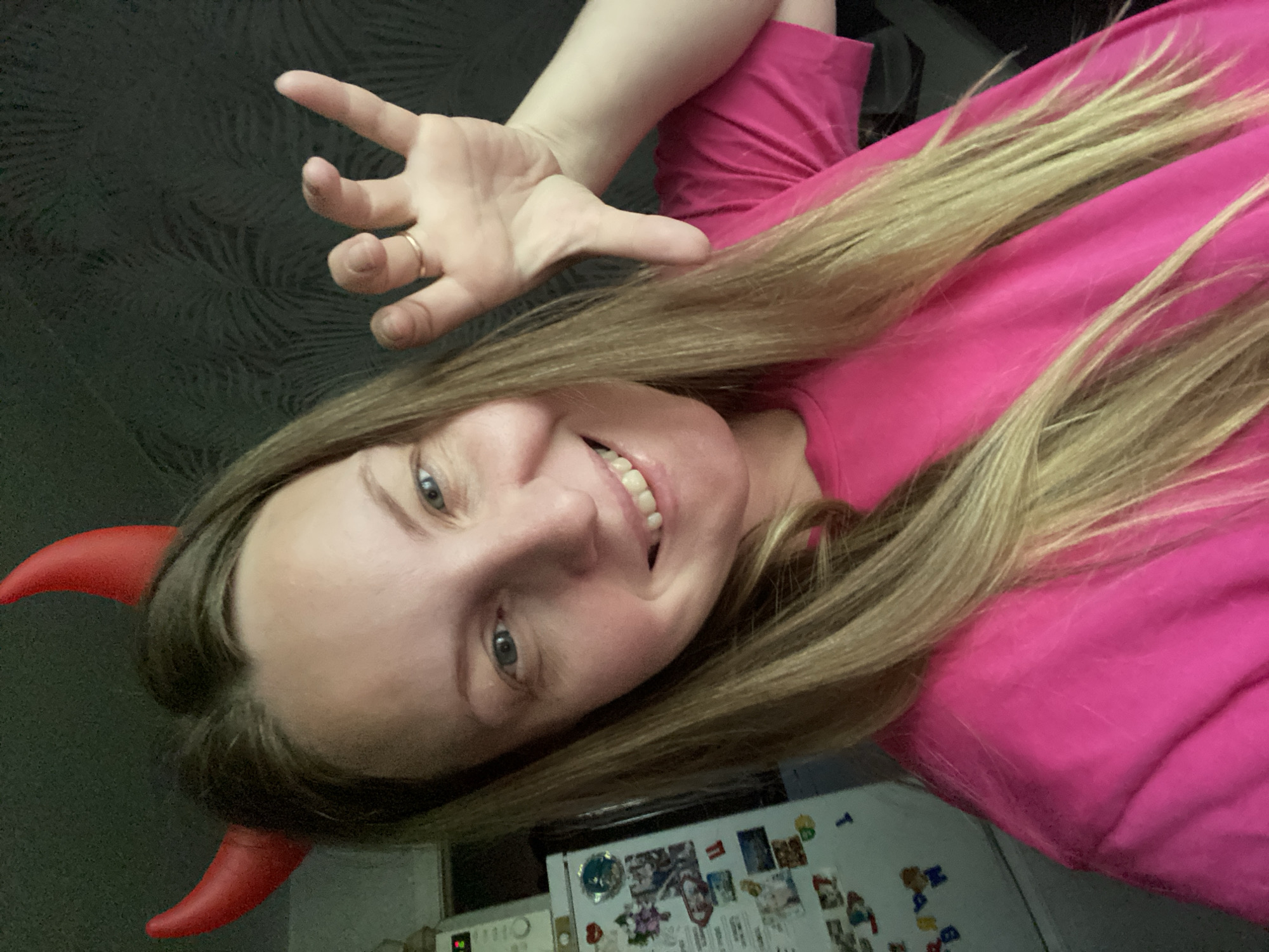 A woman with long blonde hair is facing the camera, wearing a pink shirt and red devil horns. She is smiling and holding her hand up with her fingers slightly spread apart. She is wearing a ring on her finger. Behind her is a wall with a patterned design. A refrigerator covered with magnets is visible in the lower right corner. The location is not discernible as being in the city of Noviy, Russia.