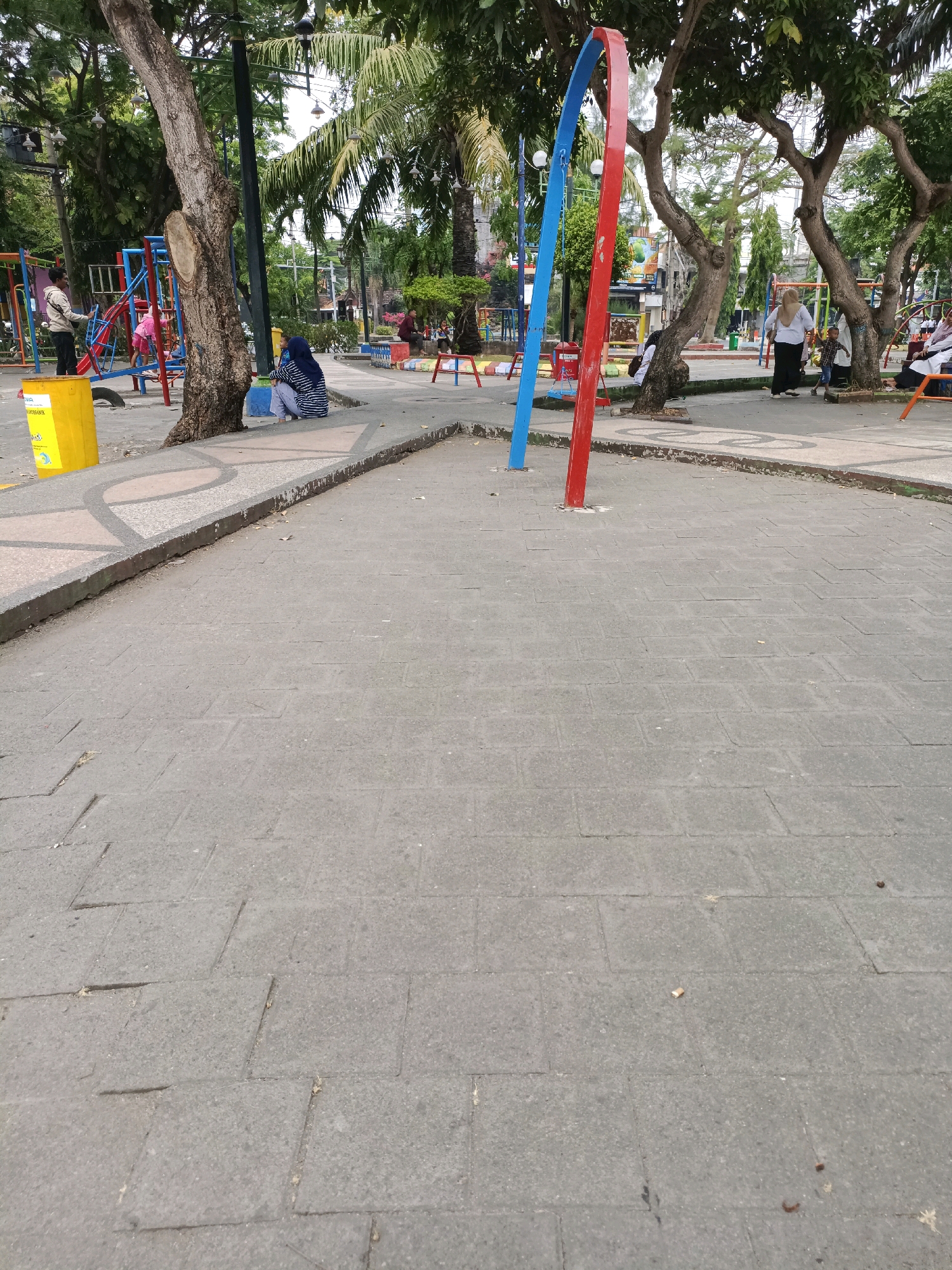 The image depicts a public park area in Lamongan, Indonesia. In the foreground, a paved walkway made of grey rectangular tiles dominates the scene. The walkway has a decorative border with a pattern of curved light-colored shapes and darker triangular accents. A low, concrete curb separates the walkway from a dirt and leaf-strewn area to the left, where a large, weathered tree stands. Beside the tree, a bright yellow trash receptacle is visible.
Further into the park, various elements of a playground are present, including red and blue metal structures, swings, and a slide. Several people are scattered throughout the park. To the left, near the playground, a person in a blue and white striped hijab sits on the ground. A man in a light-colored jacket stands near the playground equipment. In the middle ground, a prominent structure with curved red and blue poles stands in the paved area. To the right of this structure, other individuals, including a woman in a white top and headscarf with a child, are present near playground equipment. In the background, more trees and some buildings are visible. The overall environment appears to be an outdoor recreational space with a mix of paved pathways and natural elements.