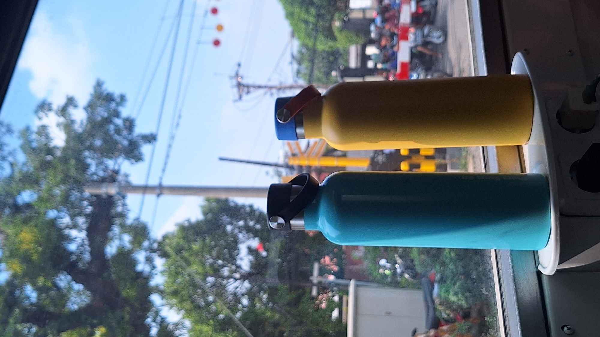 Two metal water bottles, one yellow and one turquoise, are inside a vehicle, likely a train or bus. They are secured in a white holding fixture attached to a window frame. Outside the window, a street scene is visible, with power lines, trees, traffic lights, and numerous motorcycles and vehicles. The location is likely in Cikarang, Indonesia, although this cannot be definitively confirmed from the image alone.