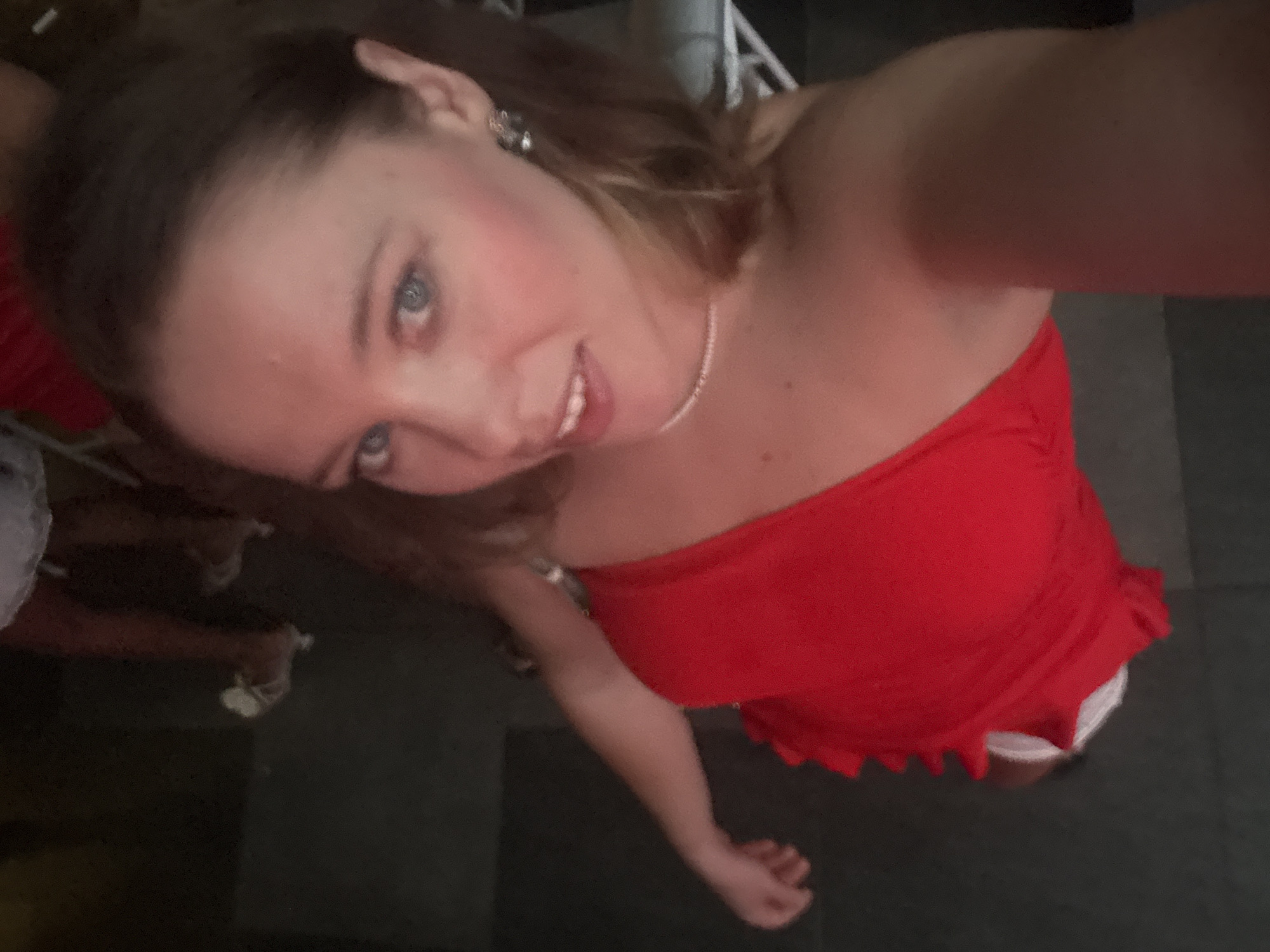 This is a selfie of a young woman with light brown hair and blue eyes, wearing a red strapless top and a pearl necklace. She is smiling, and her head is tilted to the side. The background is dark and appears to be an indoor setting, possibly a room or a club. There are some blurry objects in the background, but they are not clearly identifiable. The lighting is dim, suggesting it might be nighttime or a dimly lit environment. The woman's skin tone is fair. The image is taken from a high angle, looking down at the subject. There is no visible text in the image. The context provided suggests the location is Đà Nẵng, Vietnam, but there are no specific visual cues to confirm this.