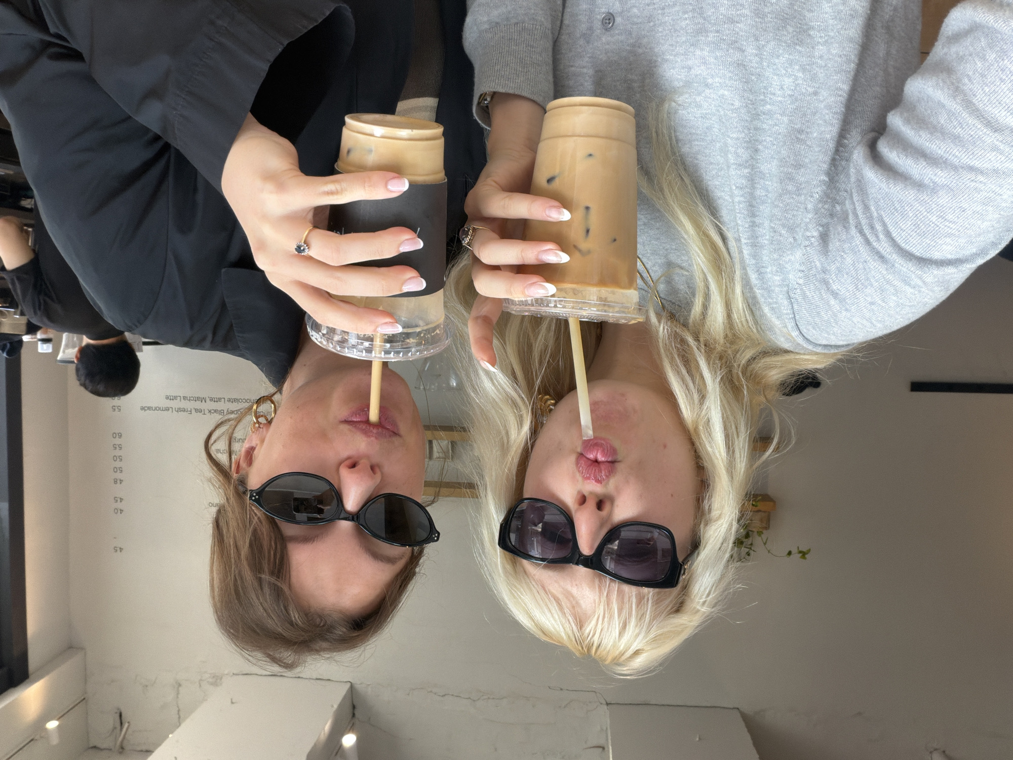 Two women wearing sunglasses are drinking from coffee cups with straws. The woman on the left has brunette hair and is wearing a black jacket. The woman on the right has blonde hair and is wearing a gray sweater. They are both wearing rings. A menu board is visible in the background. The image is inverted. Based on the "purg" menu item on the board, the image was likely taken at Purg in Seoul, South Korea.