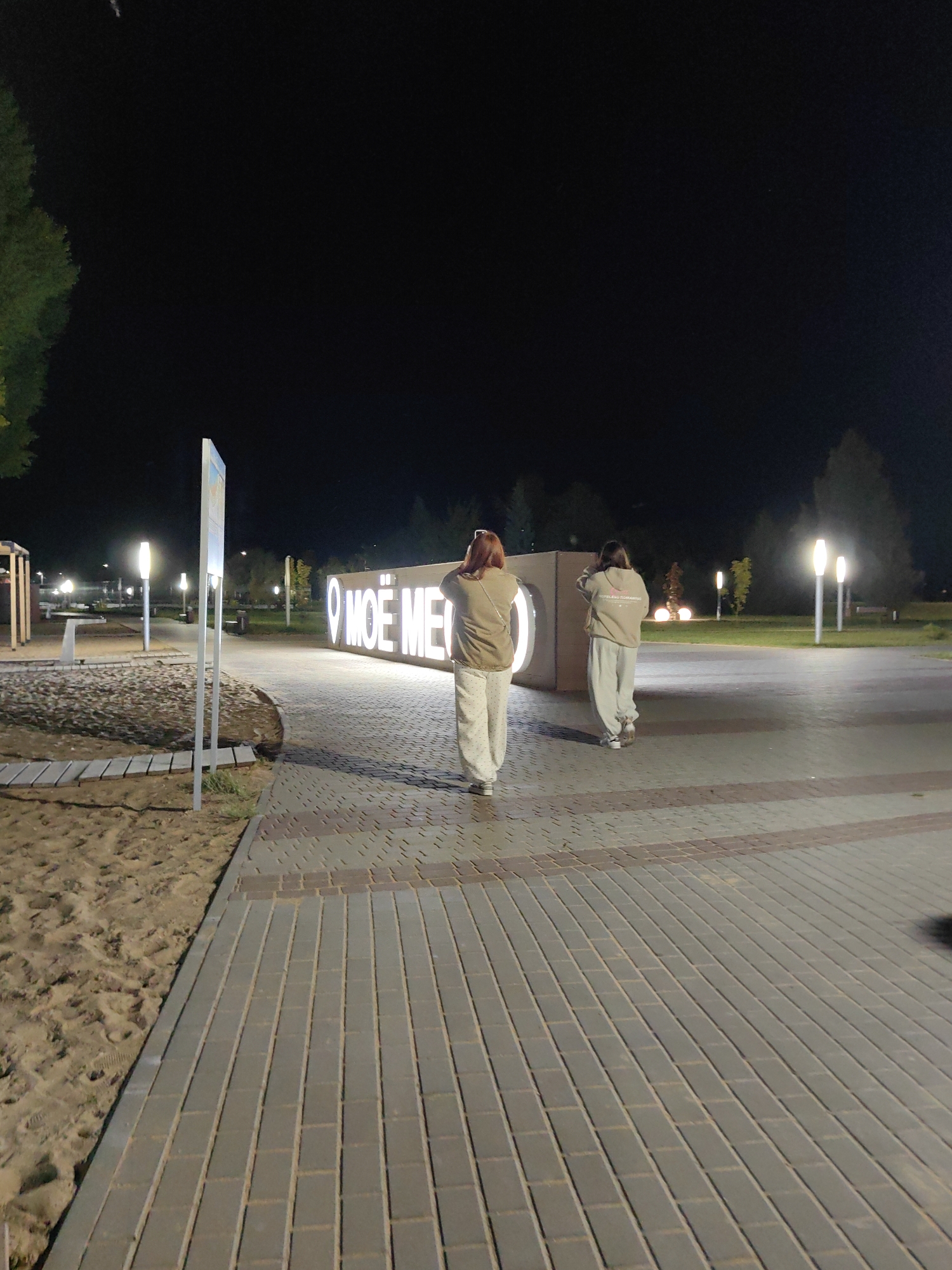 Two young women walk away from the camera on a paved pathway at night. The pathway is made of grey bricks arranged in a herringbone pattern, with a strip of reddish-brown bricks interspersed. To the left of the pathway is a sandy area with some wooden planks laid down, possibly a children's play area. In the background, a large illuminated sign reads "МОЁ МЕ" in white block letters. Several lampposts with warm, glowing lights are visible along the pathway and in the grassy area beyond. Trees can be seen in the background, and the sky is dark. The overall impression is of a public space at night, possibly a park or promenade. This location is in the city of Myadzyel, Belarus.