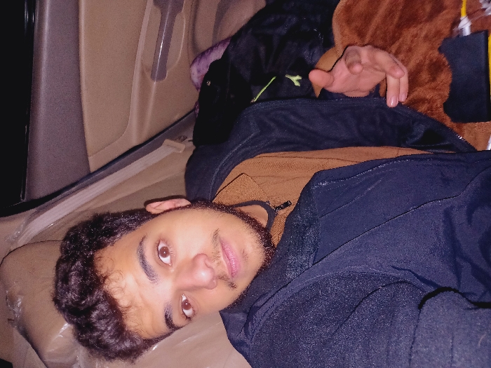 A young man with curly dark hair, brown eyes, and a beard lies on a beige surface, seemingly inside a vehicle. He is wearing a brown sweater and a black jacket. Above him is a mix of items, including a black backpack with a green logo and a brown, fuzzy object. The vehicle's interior features a door panel with a handle. The overall lighting is dim.