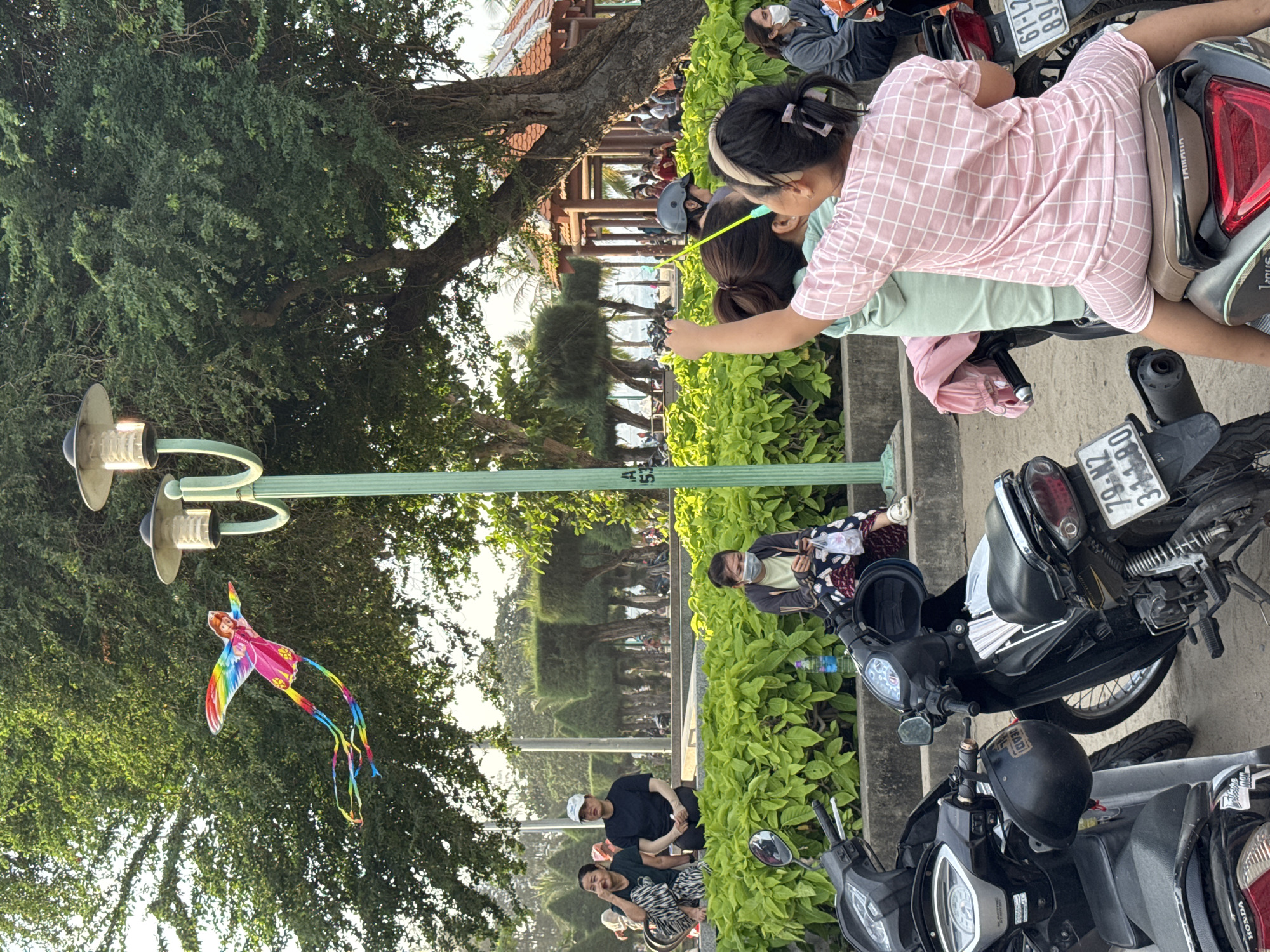 The image captures a scene in a public park in Nha Trang, Vietnam, likely near the beach given the presence of palm trees and a glimpse of the ocean in the background. In the foreground, several motorcycles are parked. A young girl wearing a pink checkered outfit is seated on one of the motorcycles, facing away from the camera. She appears to be holding a thin, yellow pole, possibly for a kite or a toy. Behind her, another person, possibly an adult, is also partially visible.
Further back, a woman in a light green shirt and patterned pants is seated on a ledge, looking at her phone. She is wearing a face mask. To her left, two women are sitting on the same ledge, one in a dark t-shirt and white cap, and the other in a dark green shirt and patterned pants. The woman in the dark green shirt is looking to her left, with her hand touching her face.
Overhead, a colorful kite with a rainbow design and a depiction of a person with wings is soaring against a backdrop of lush green trees and a clear sky. The kite is positioned near a lamppost. The overall environment suggests a relaxed outdoor setting, with people enjoying a day in the park. The architecture of a building with a red tiled roof and open-air structure is visible in the mid-ground.