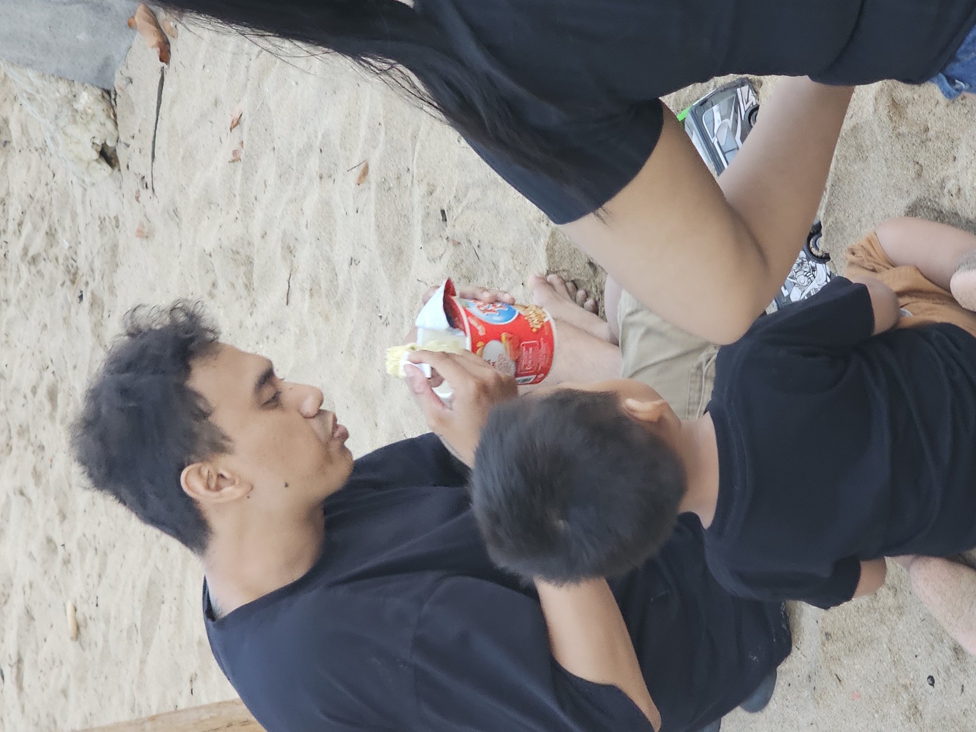 The image depicts a person with dark hair eating from a red and white container with a spoon. A child is partially visible, wearing a black shirt. Another person with long black hair and wearing a black top is also in the frame. They are situated on a sandy surface. Given the prompt, the location is presumed to be a beach or sandy area in Denpasar, Indonesia.